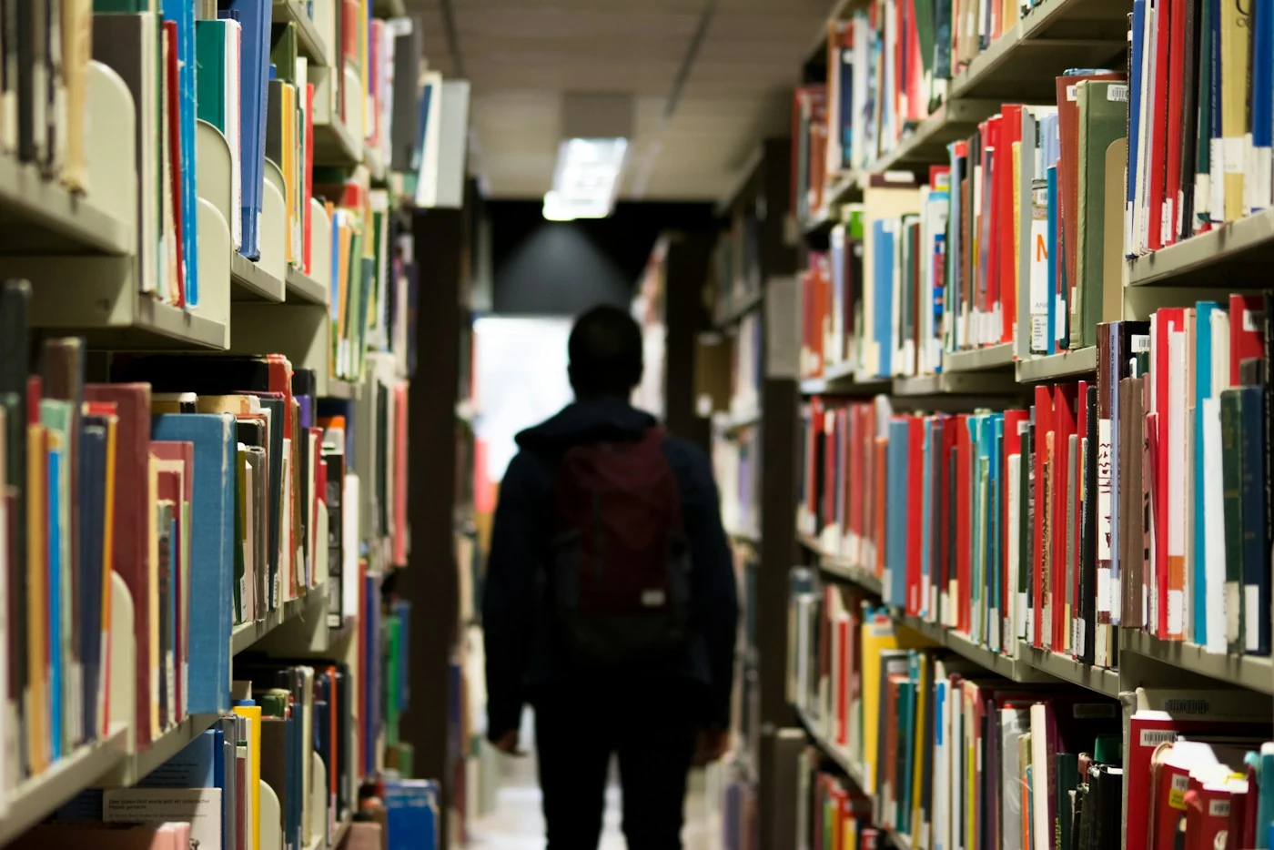 Rows of library shelves stacked with books, a reader walking away down the aisle