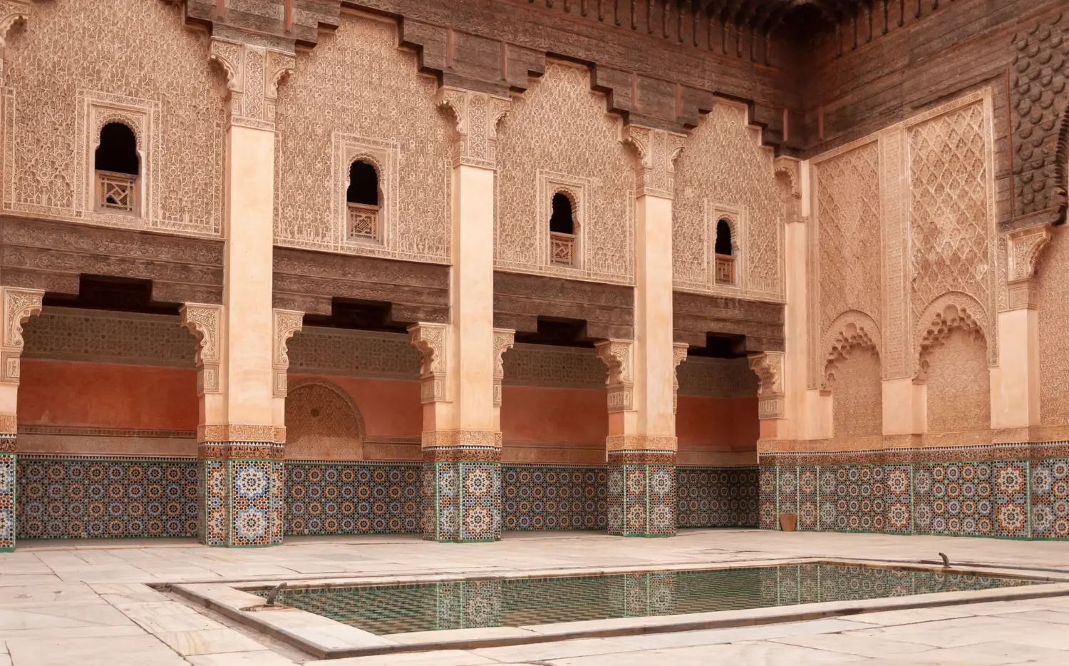 An empty courtyard of carved plaster, cedar and patterned tilework around a still reflecting pool