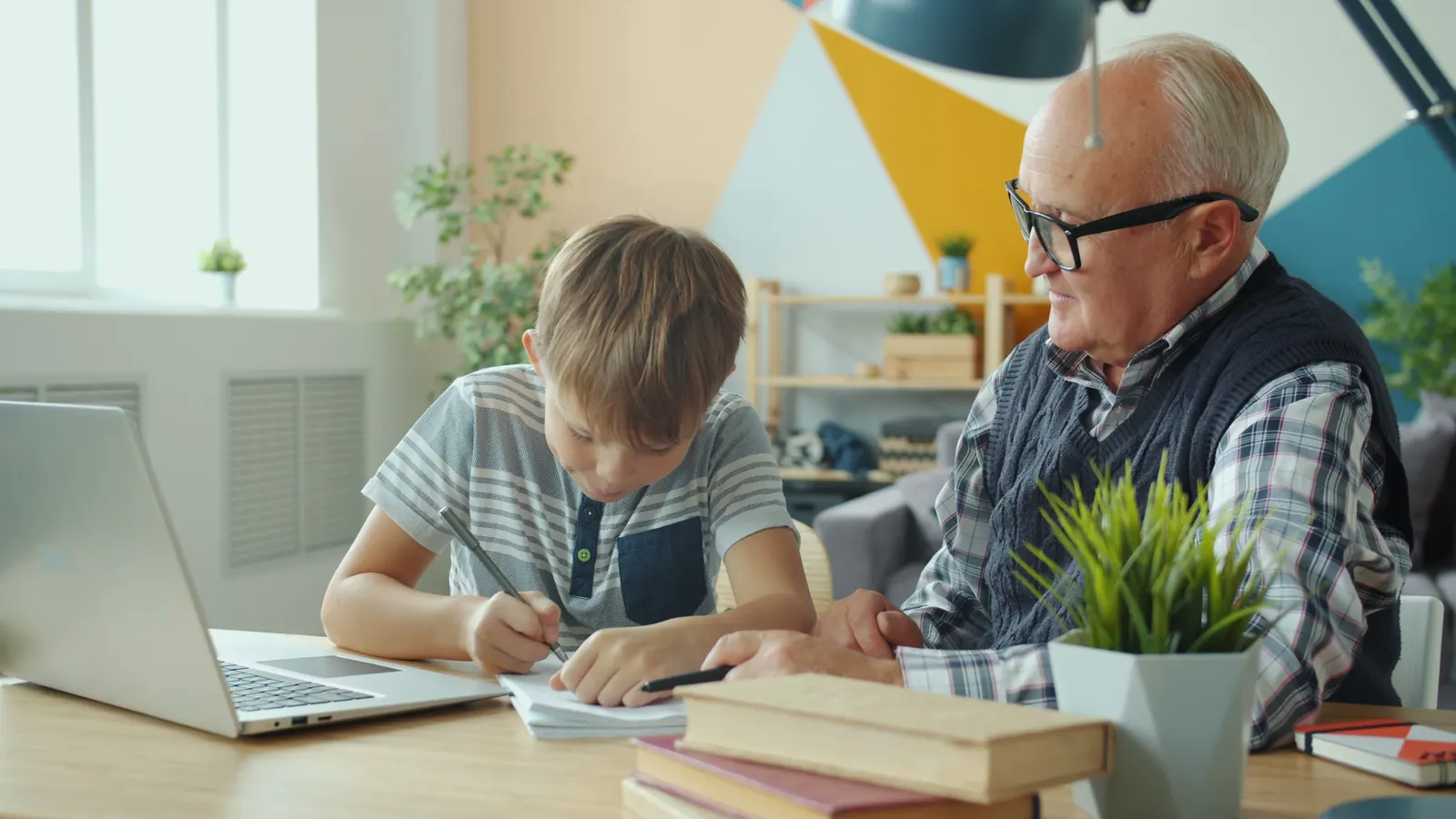 An older man in glasses sits beside a boy at a desk while the boy writes in his notebook