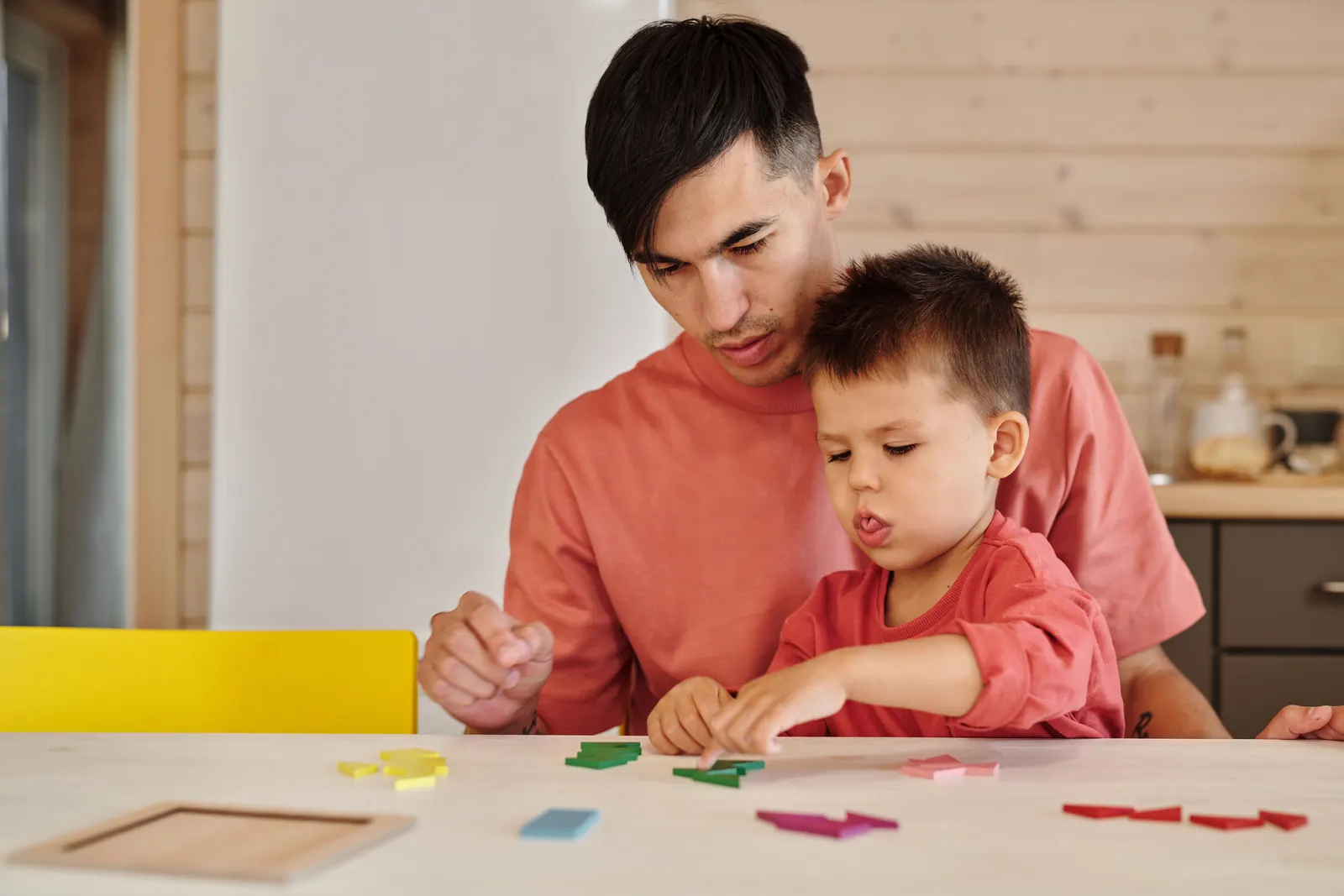 A young man sits behind a small boy at a table and guides him as he fits colored tangram pieces
