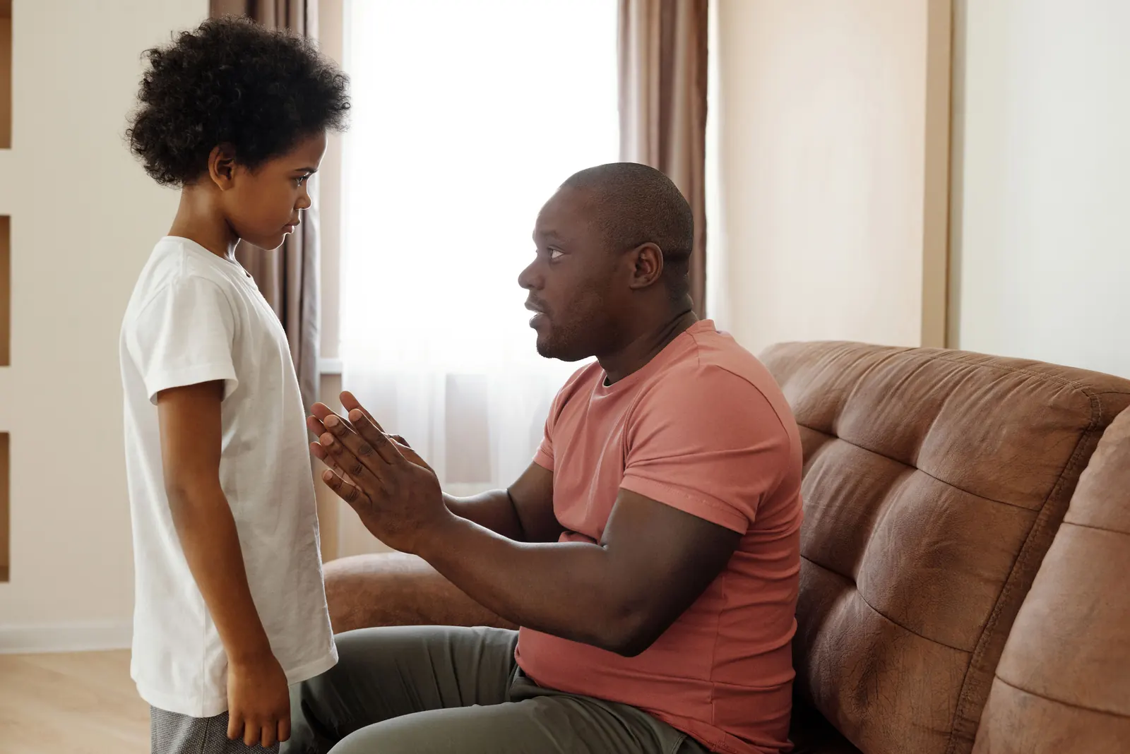 A man sitting on a leather sofa talks gently with a boy standing in front of him