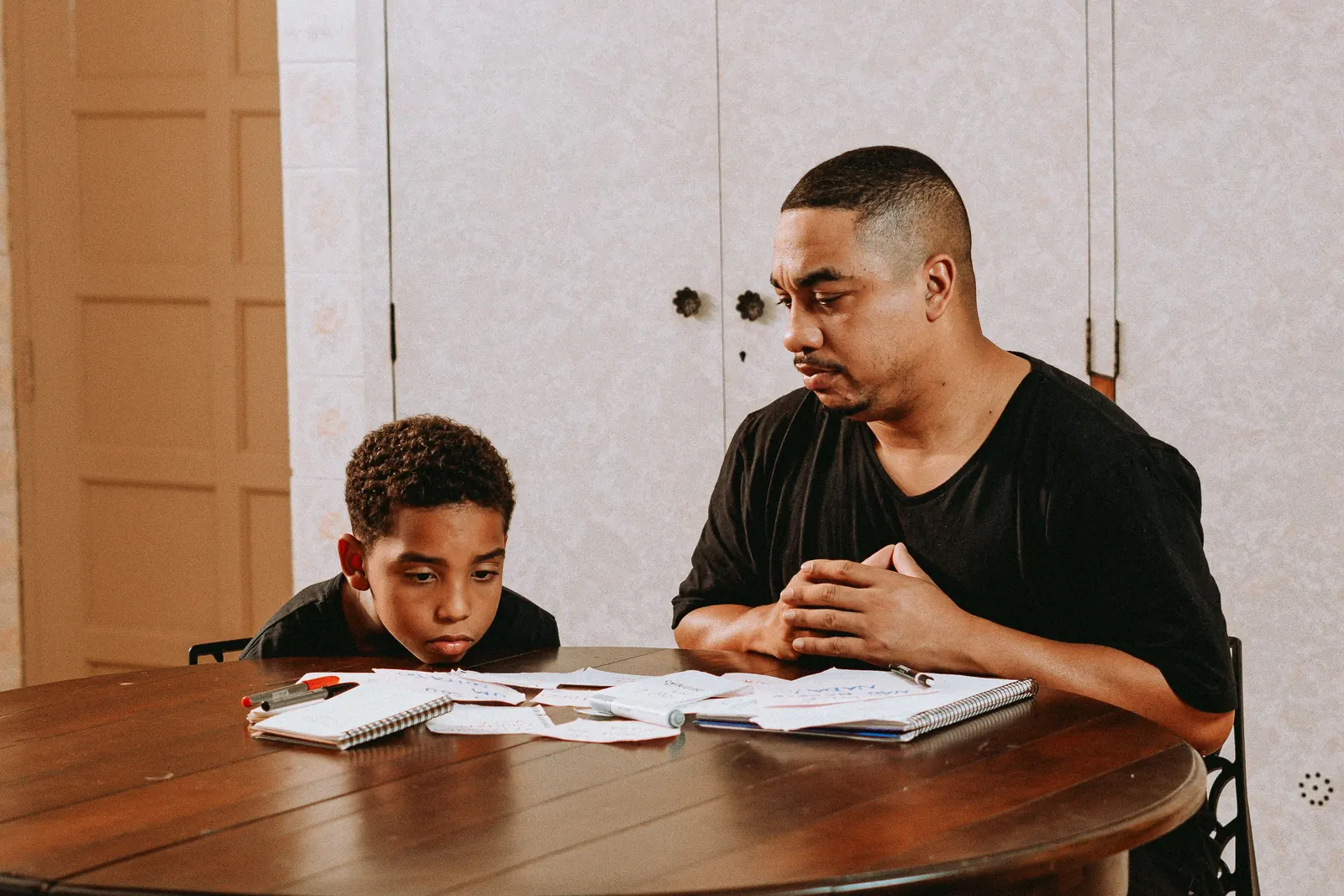A smiling man and a boy laugh together over papers at a wooden table