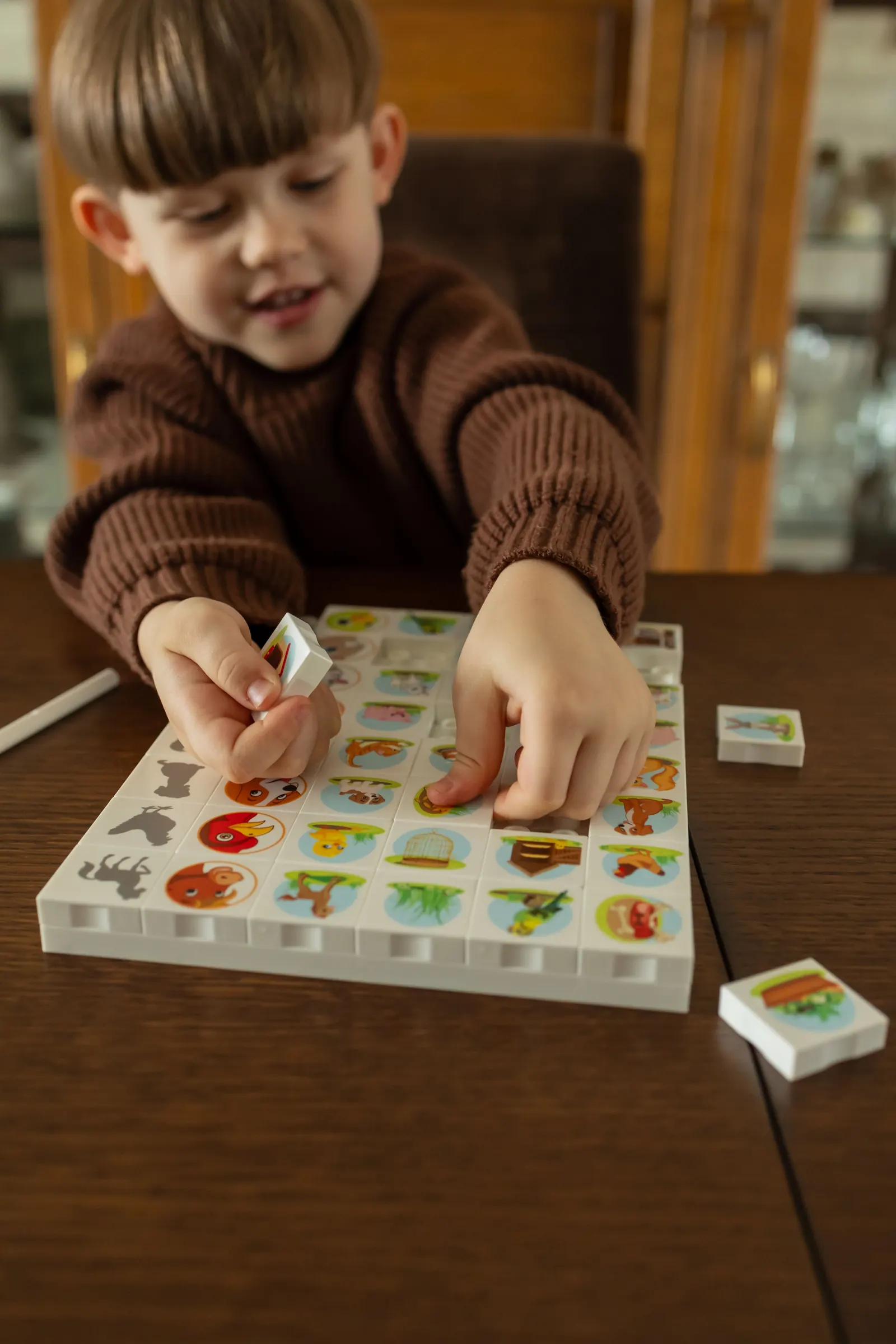 A small boy in a brown sweater matches picture tiles into a board at a wooden table