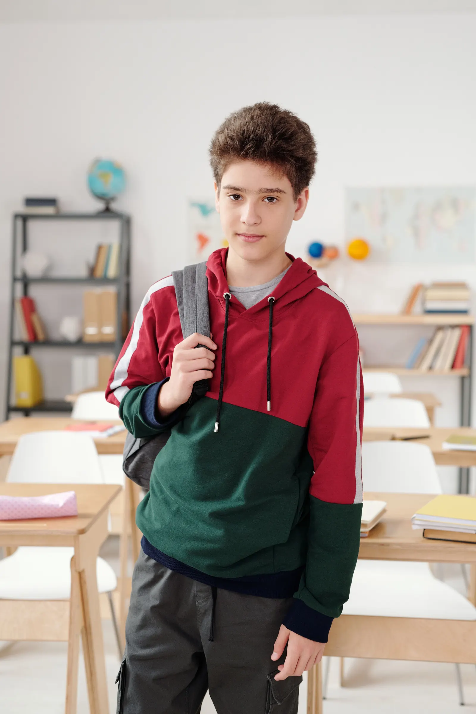 A teenage boy in a red and green hoodie stands with his backpack over one shoulder in a bright classroom