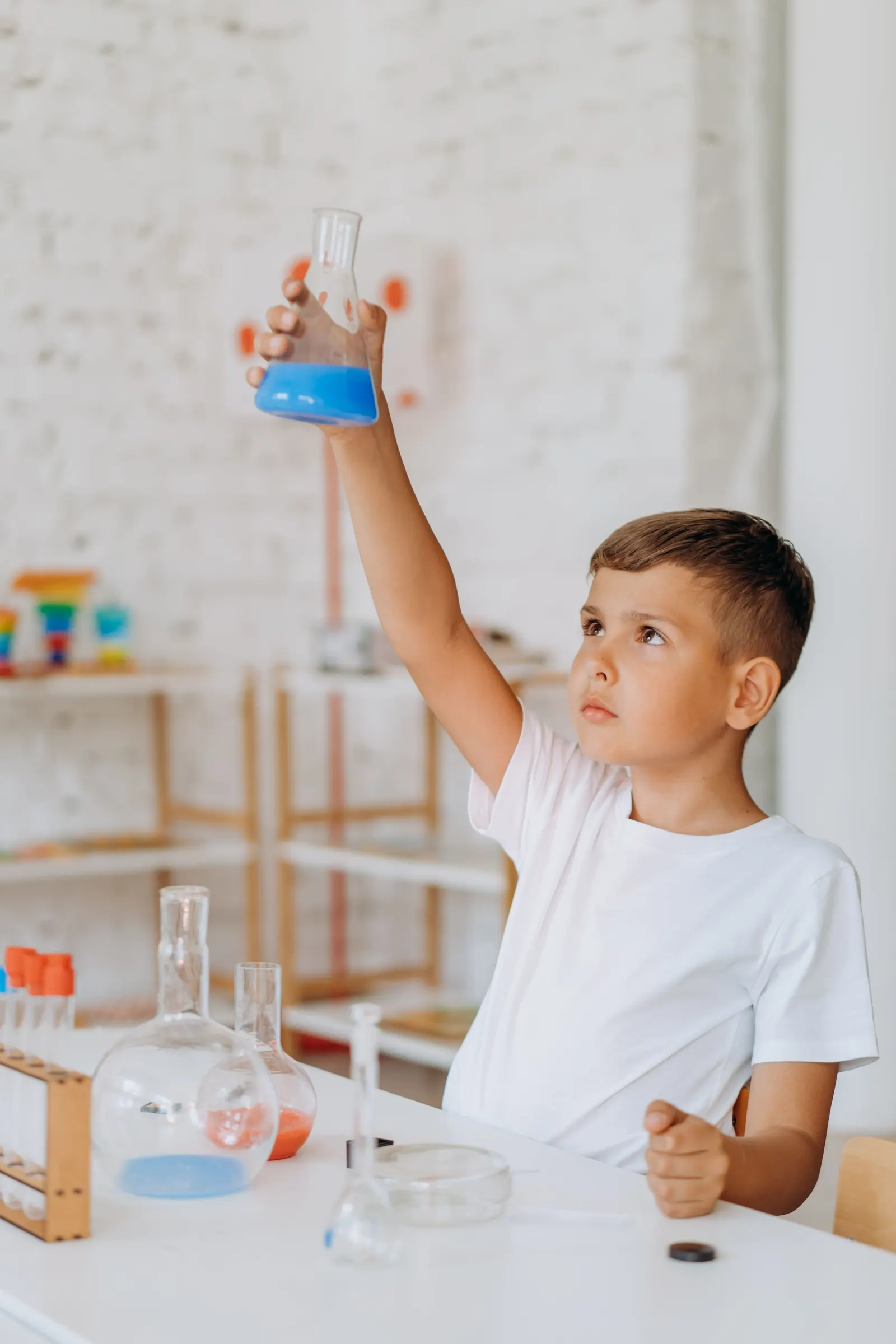 A boy in a white T-shirt holds up a flask of blue liquid, studying it, beside glass flasks and a test-tube rack