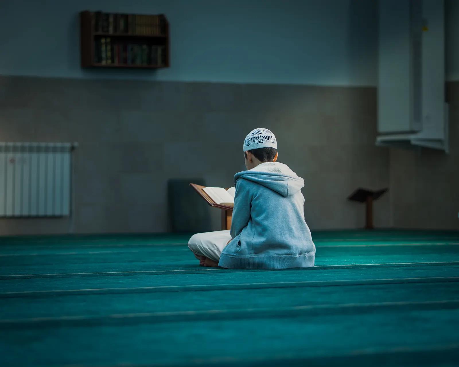 A boy in a white kufi and gray hoodie sits cross-legged on a teal mosque carpet reading from a wooden stand