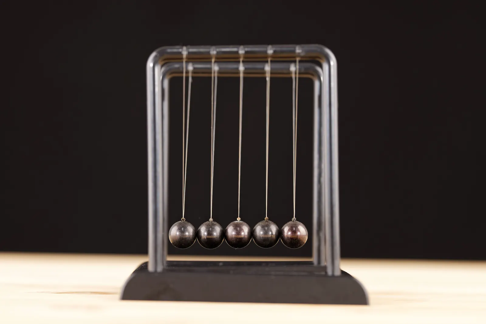 A steel Newton's cradle with five dark balls on a black base, set on light wood against a dark backdrop