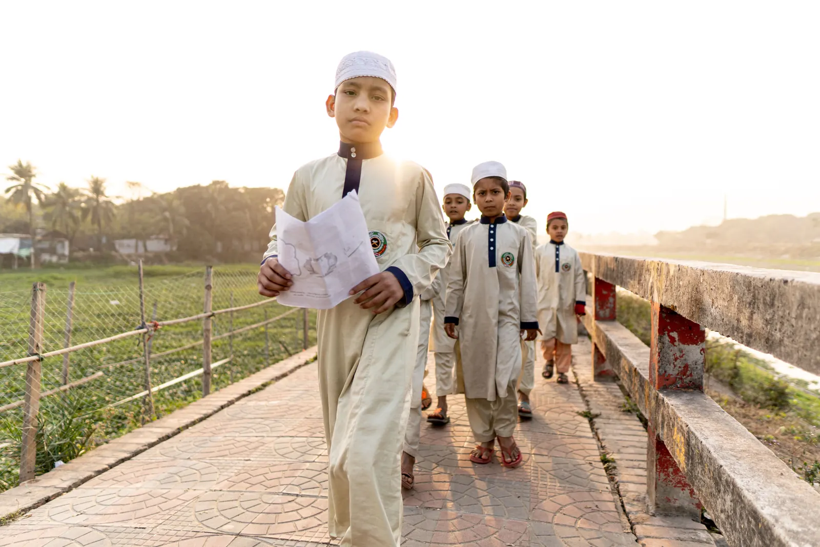 Boys in cream kurtas and white prayer caps walk in a line along a brick path beside green fields in golden light