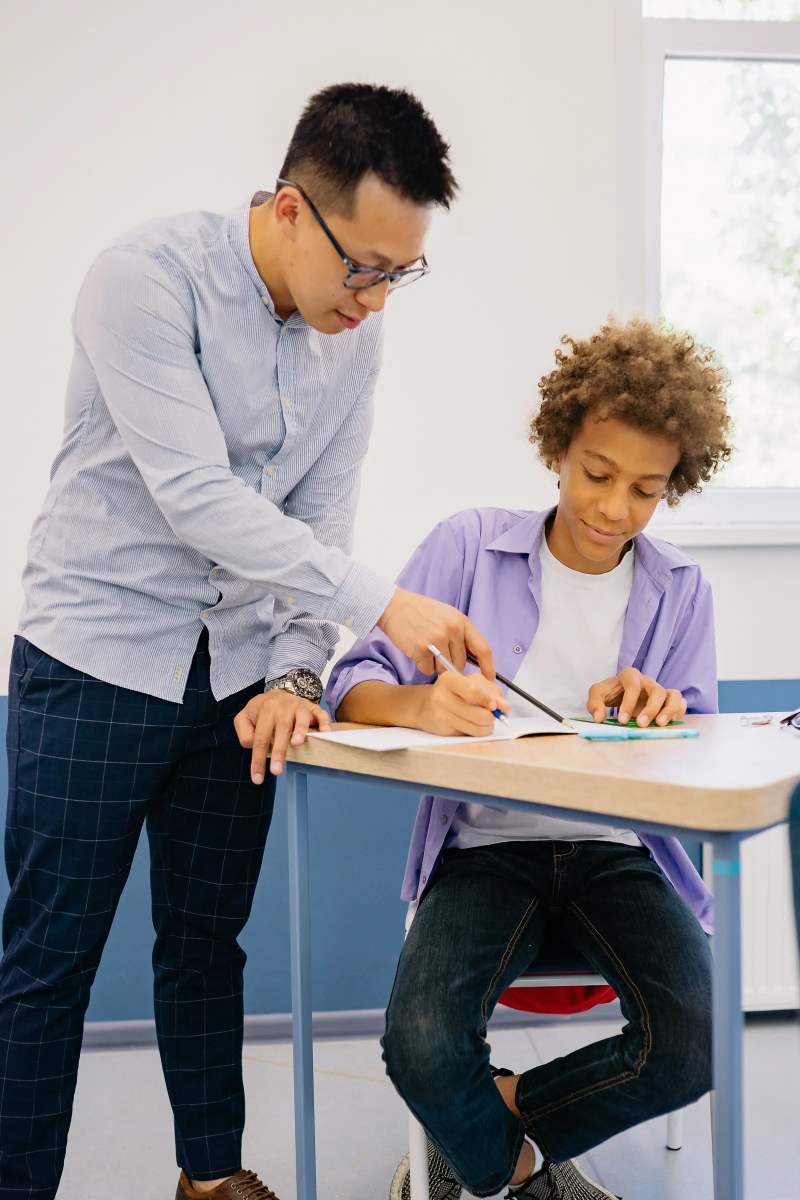 A male teacher in glasses leans over a desk to point at a boy&rsquo;s work as the boy writes