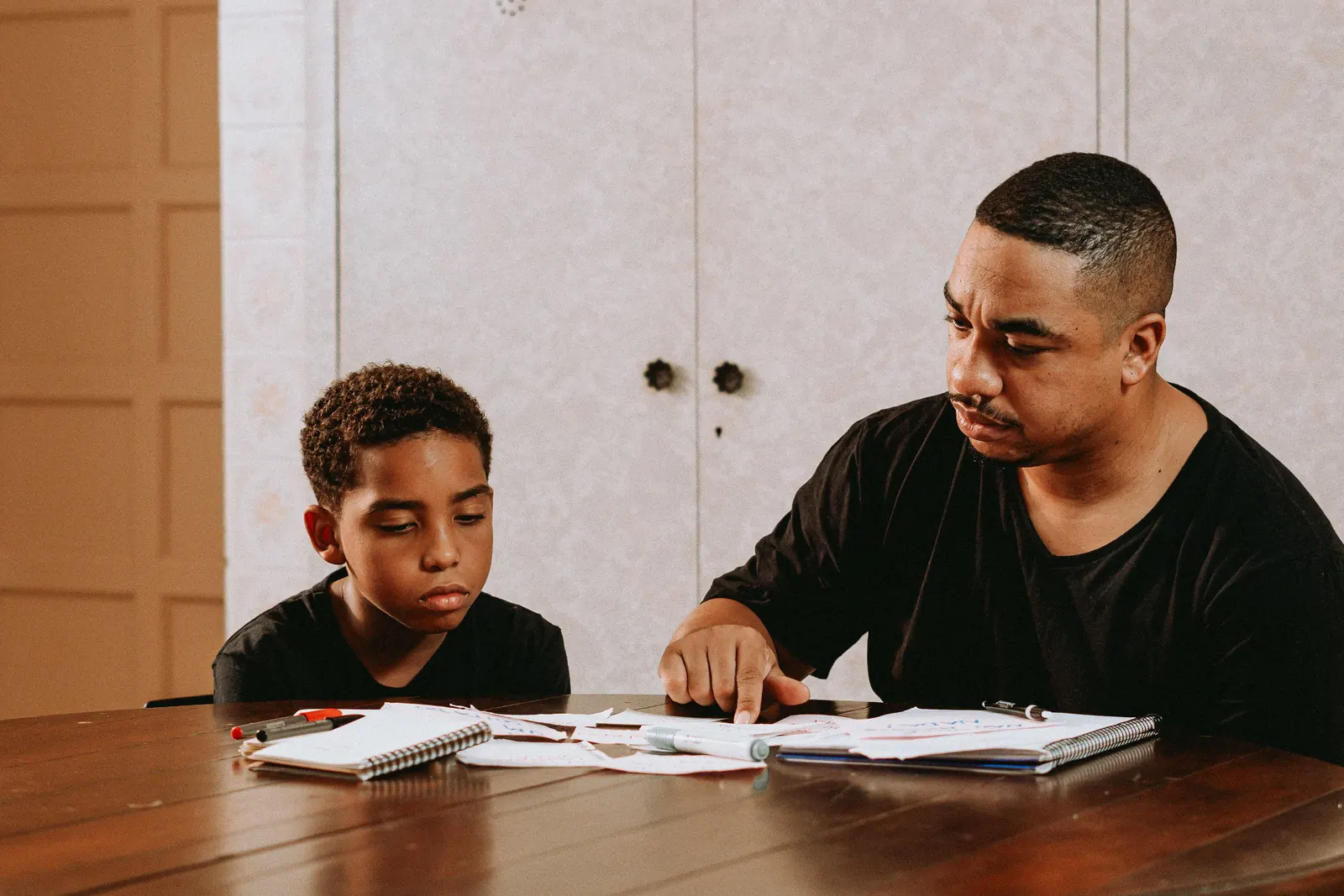 A man in a black T-shirt points to a line in a boy's notebook as they sit together at a dark wooden table