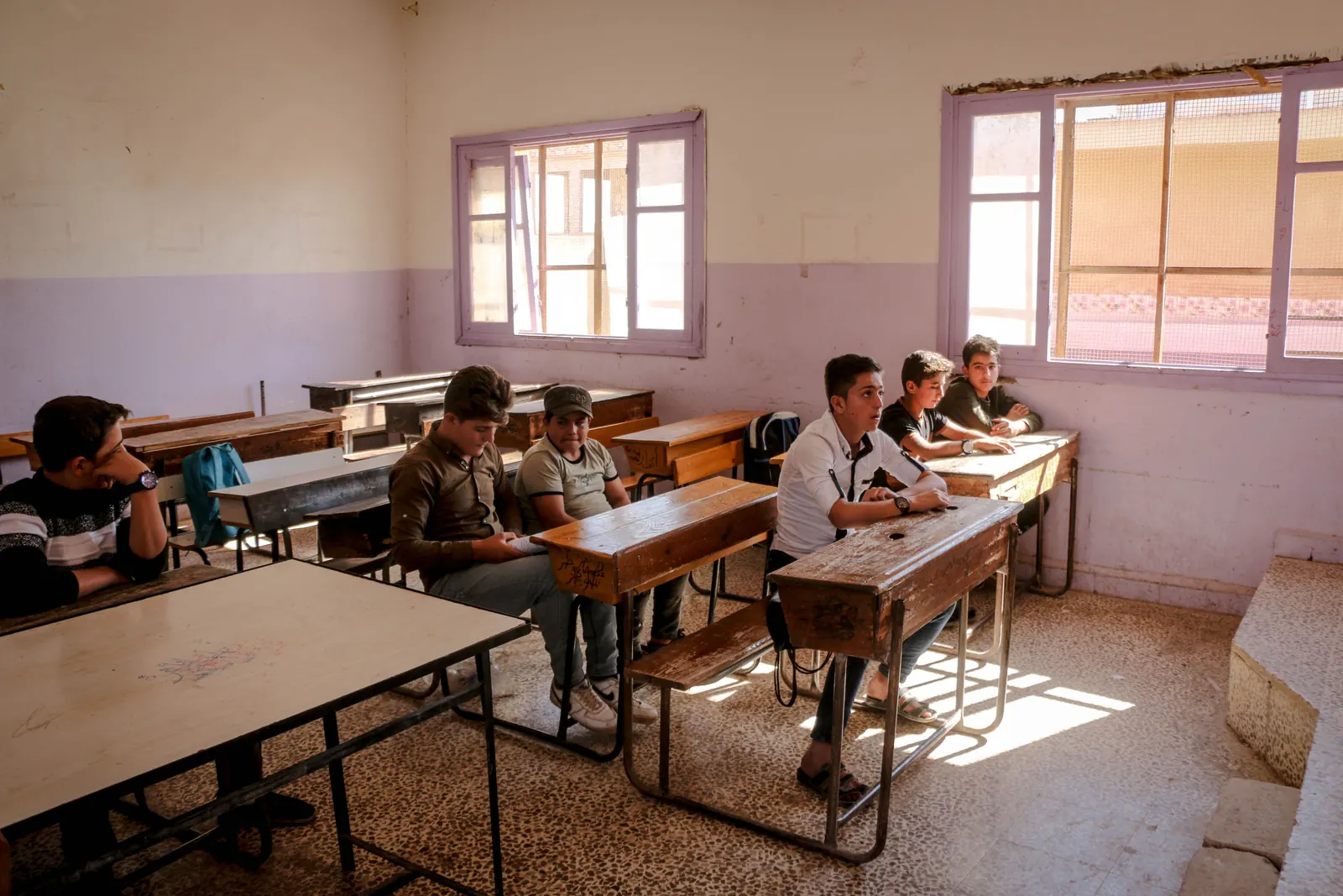 A handful of boys sit at old wooden desks in a sunlit classroom with lilac walls