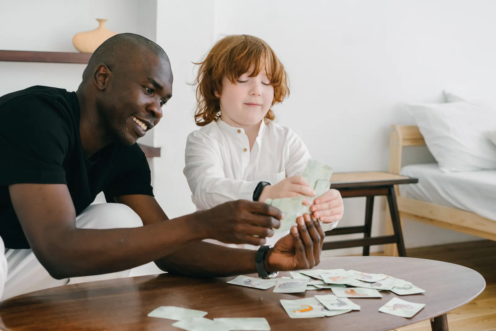 A smiling man and a red-haired boy sort picture cards together at a low wooden table