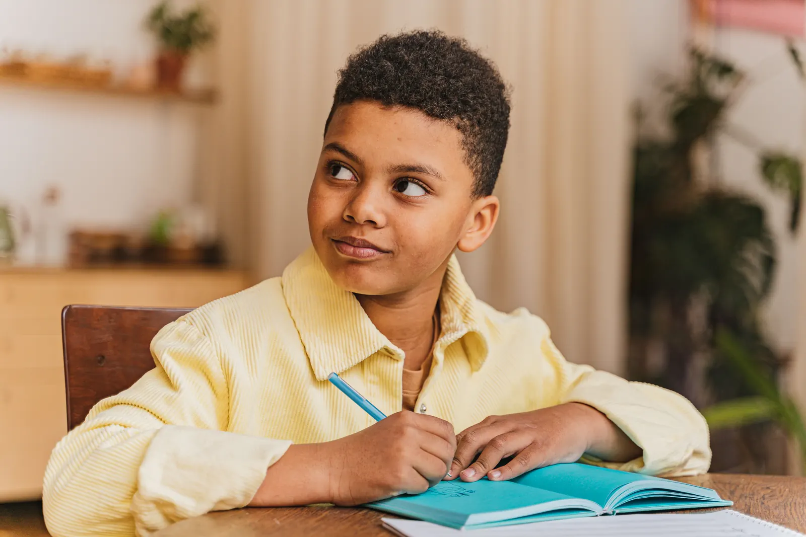 A boy in a yellow shirt pauses while writing in a notebook at a wooden table