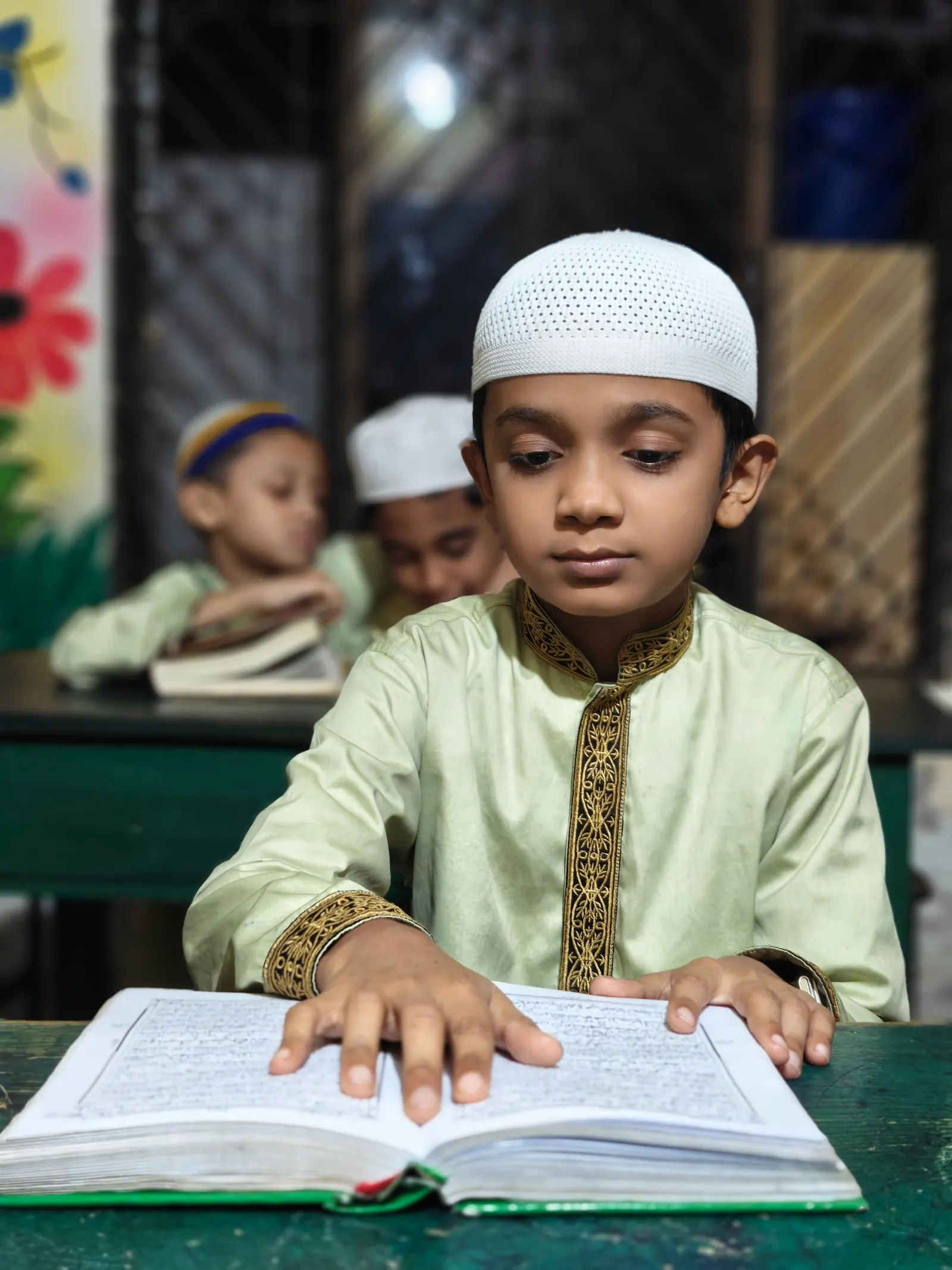 A boy in a white kufi reads from a large mushaf at a green classroom desk