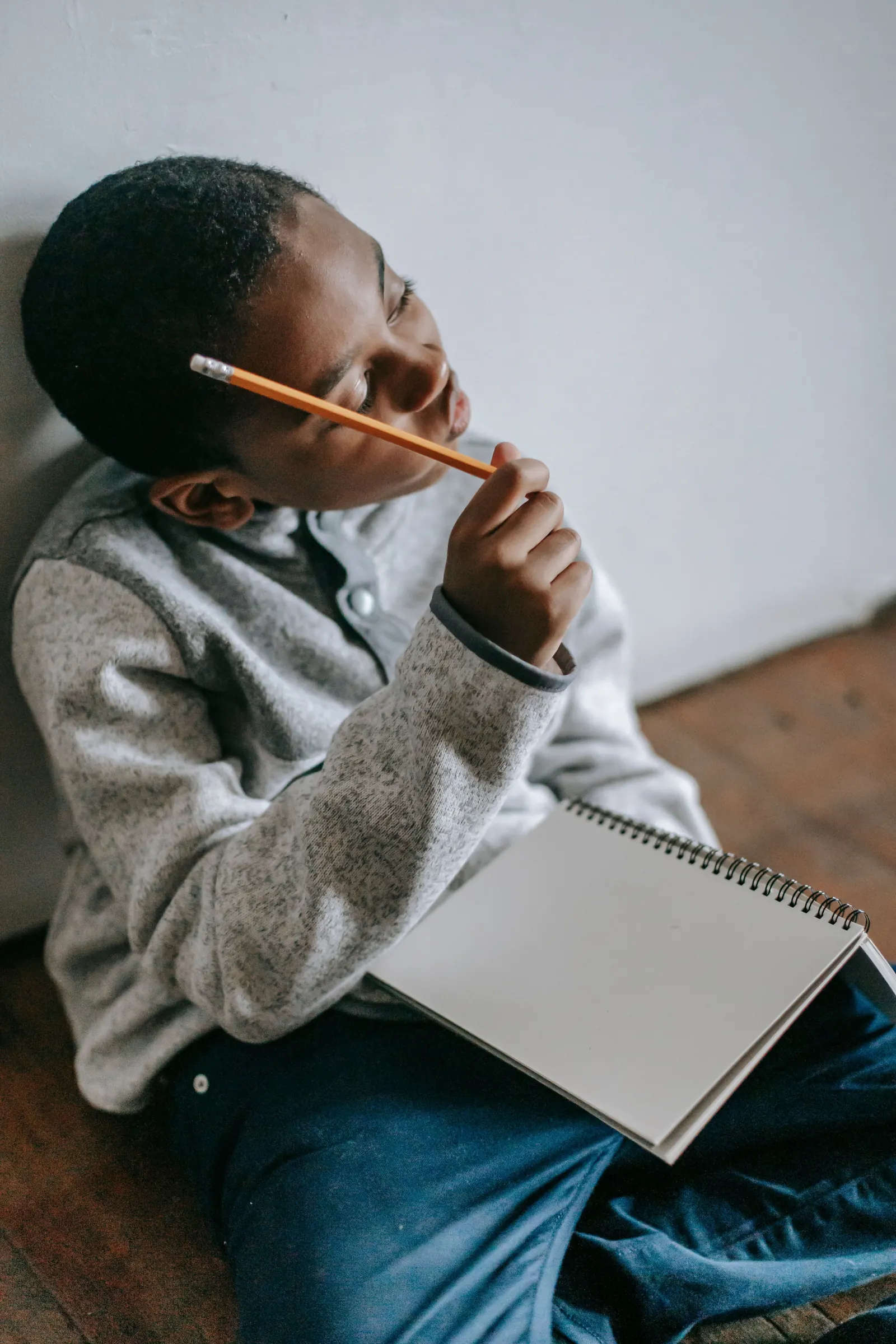 A boy sits against a wall with a sketchbook, pencil to his temple, thinking