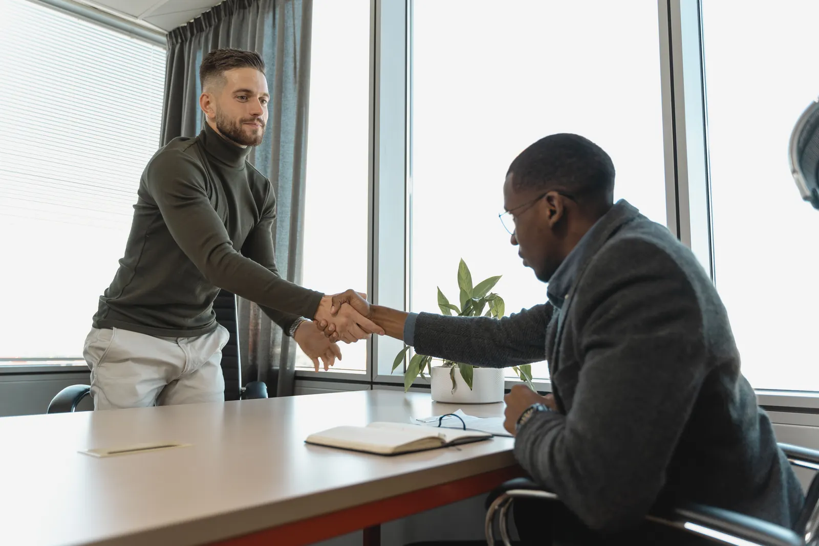 Two men shake hands across an office desk by a bright window, a notebook between them