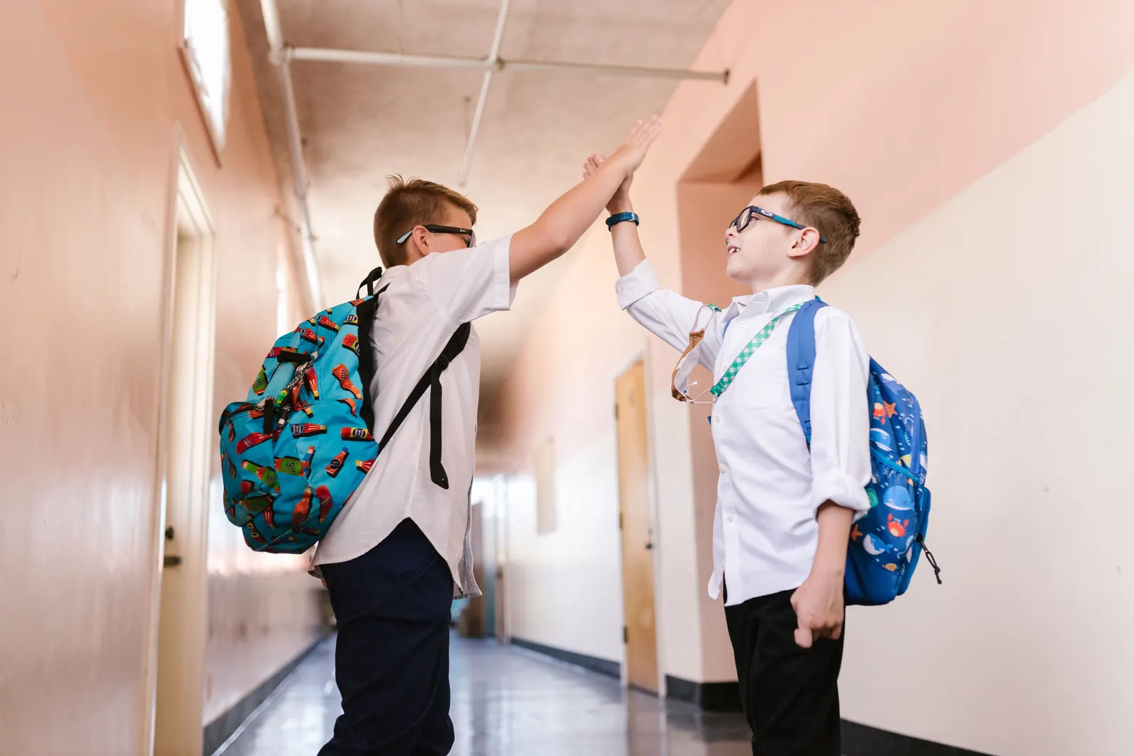 Two schoolboys in white shirts with backpacks share a high five in a sunlit school corridor