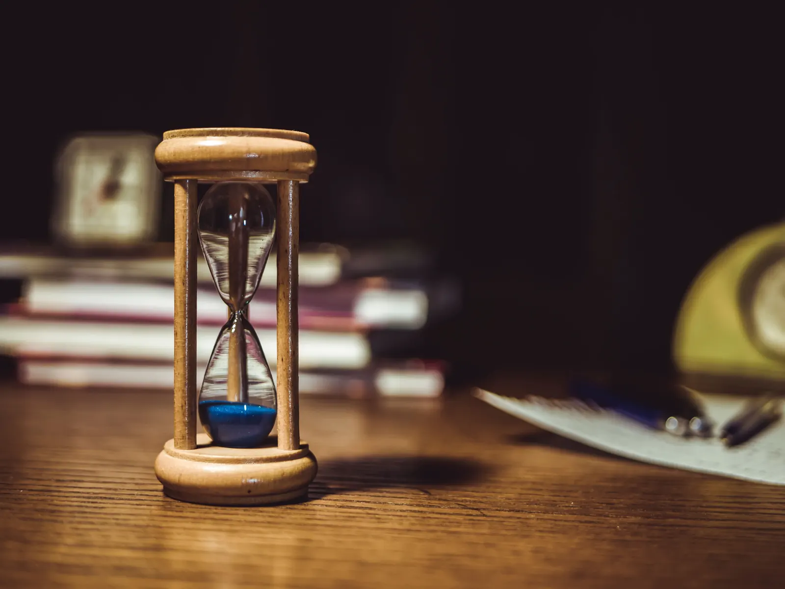 A wooden hourglass with blue sand on a warm wooden desk, books softly blurred behind
