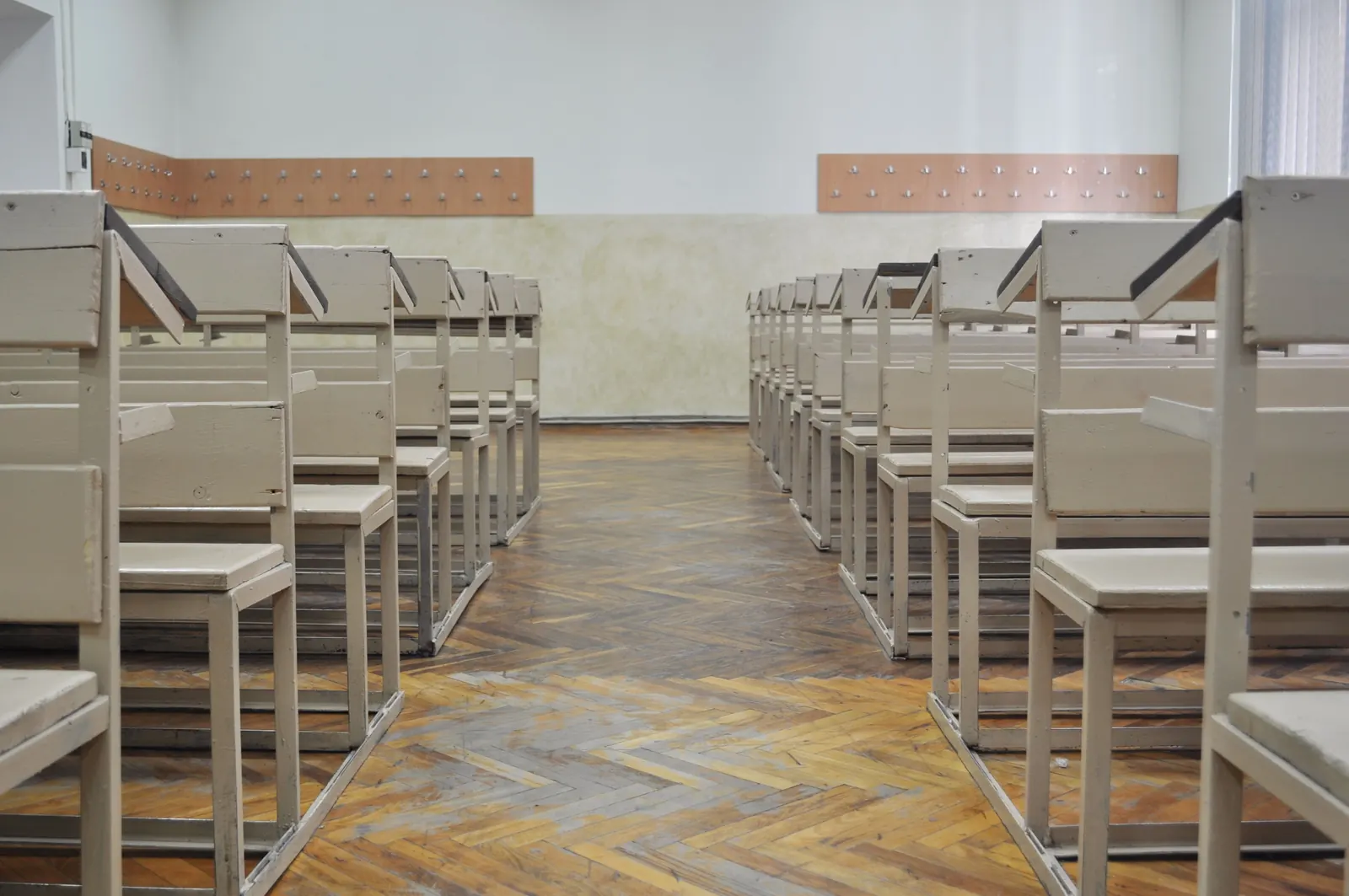 An empty classroom of old wooden desk-benches in two rows either side of a warm herringbone parquet aisle