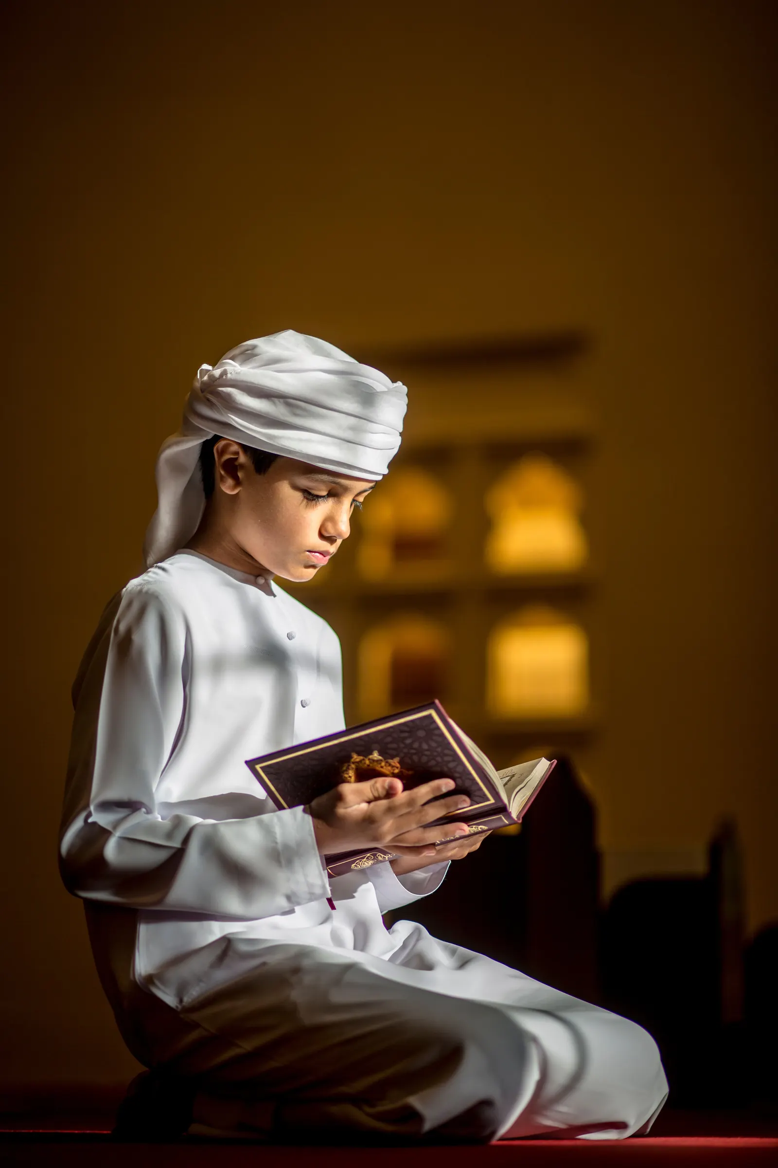 A boy in a white thobe and ghutra kneels on a prayer carpet reading the Qur'an in a warmly lit masjid
