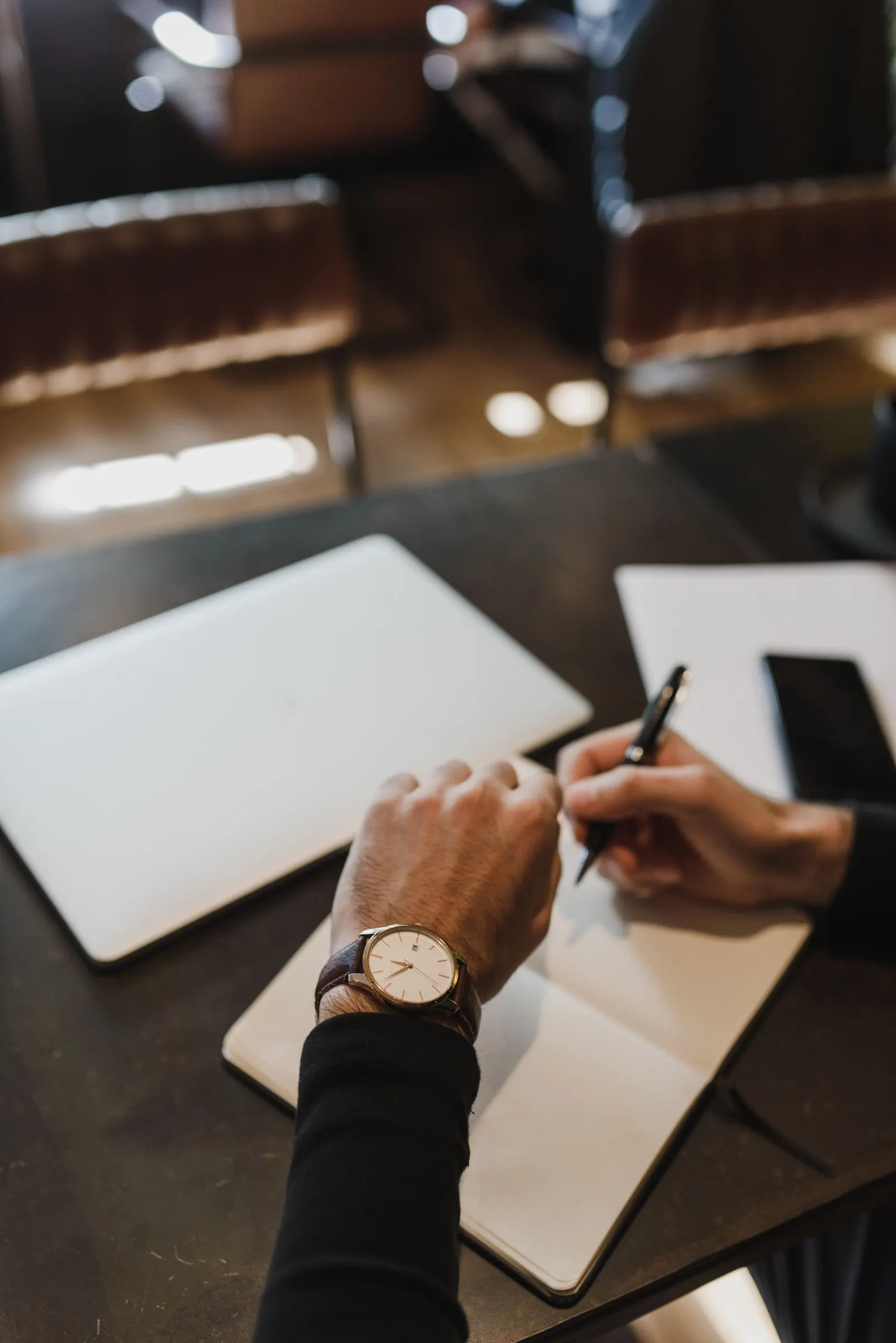A man's hands, a watch on one wrist, writing with a pen in a plain open notebook on a dark table