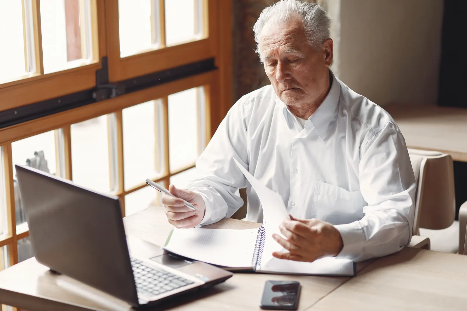 An older man in a white shirt reads through papers carefully at a desk by a sunny window