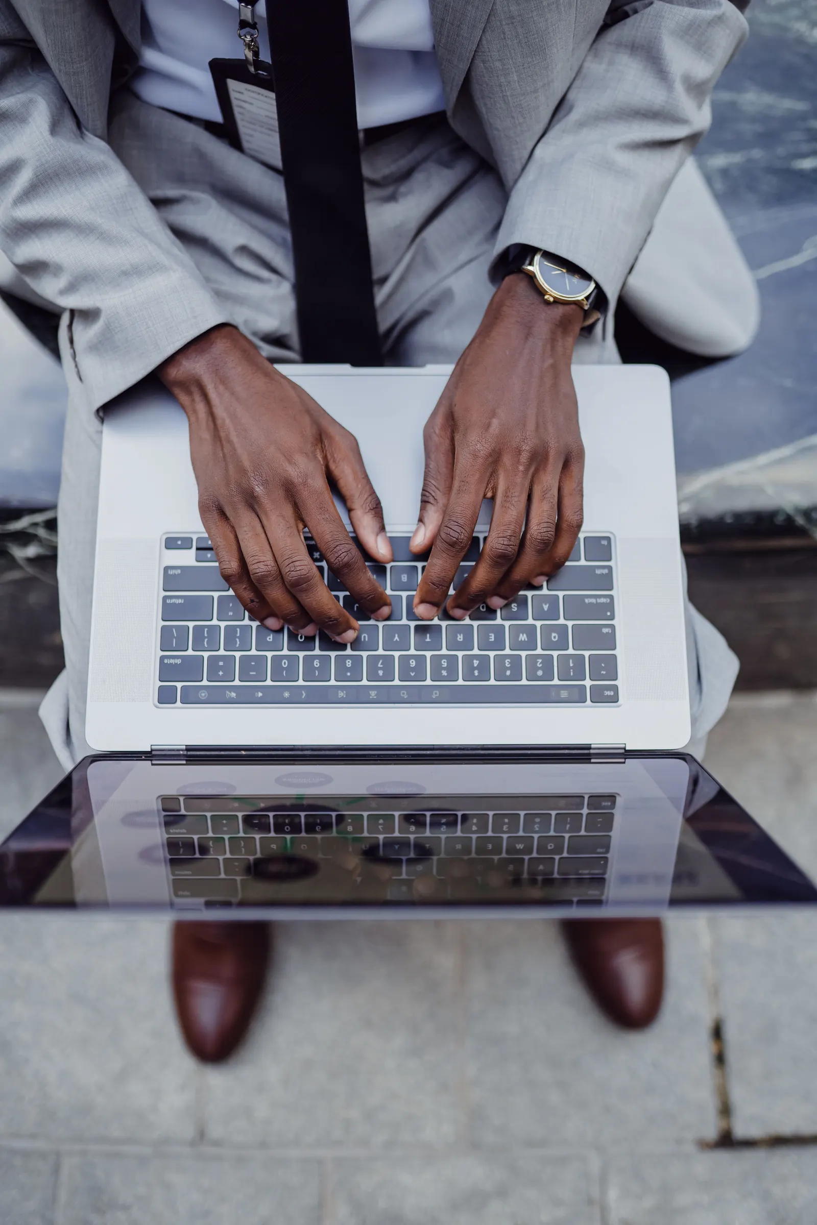 From above, a man in a grey suit types on a laptop resting on his knees
