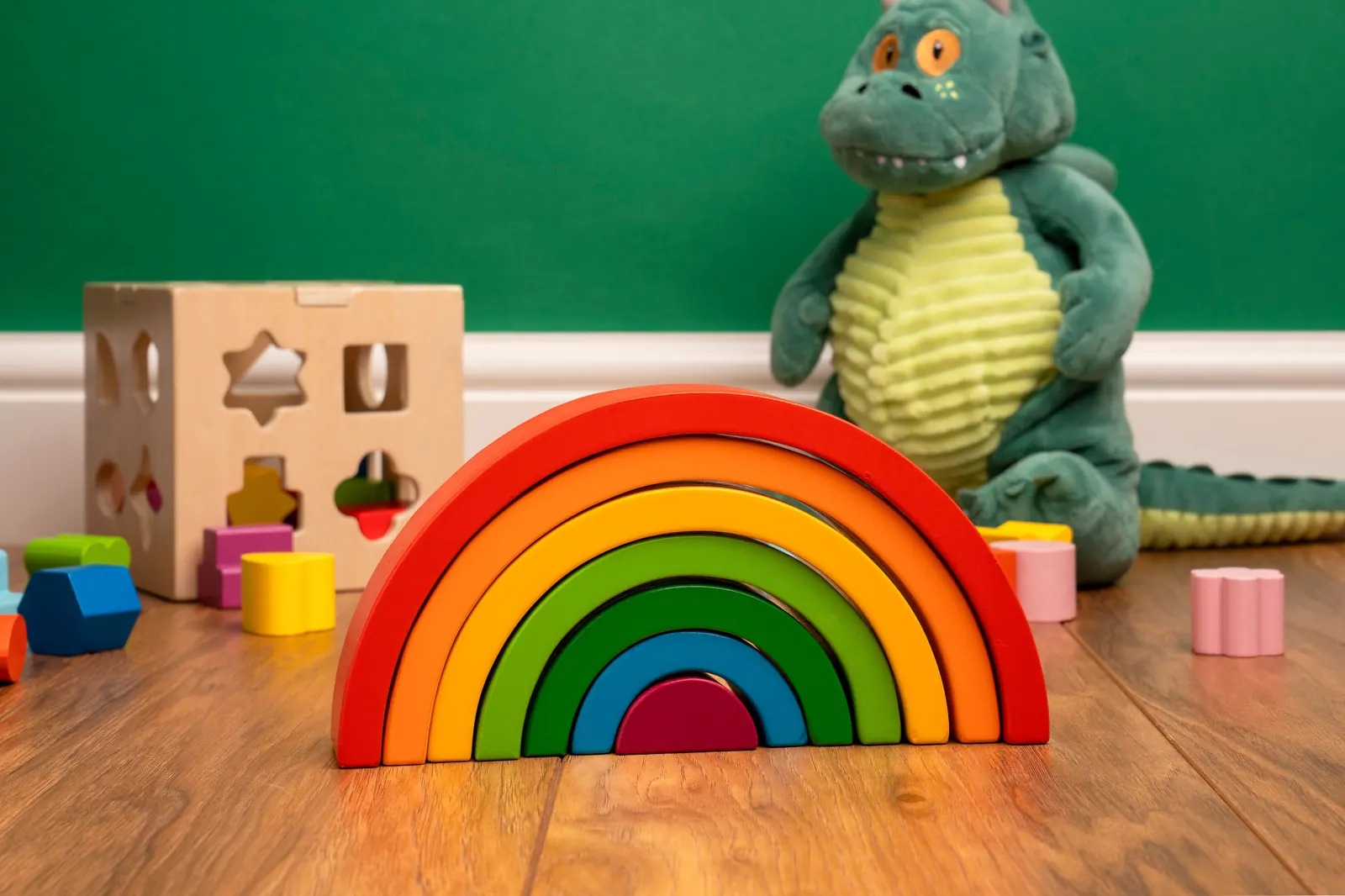 A wooden rainbow stacking toy, a wooden shape-sorter box and a soft toy dinosaur on a warm wooden floor