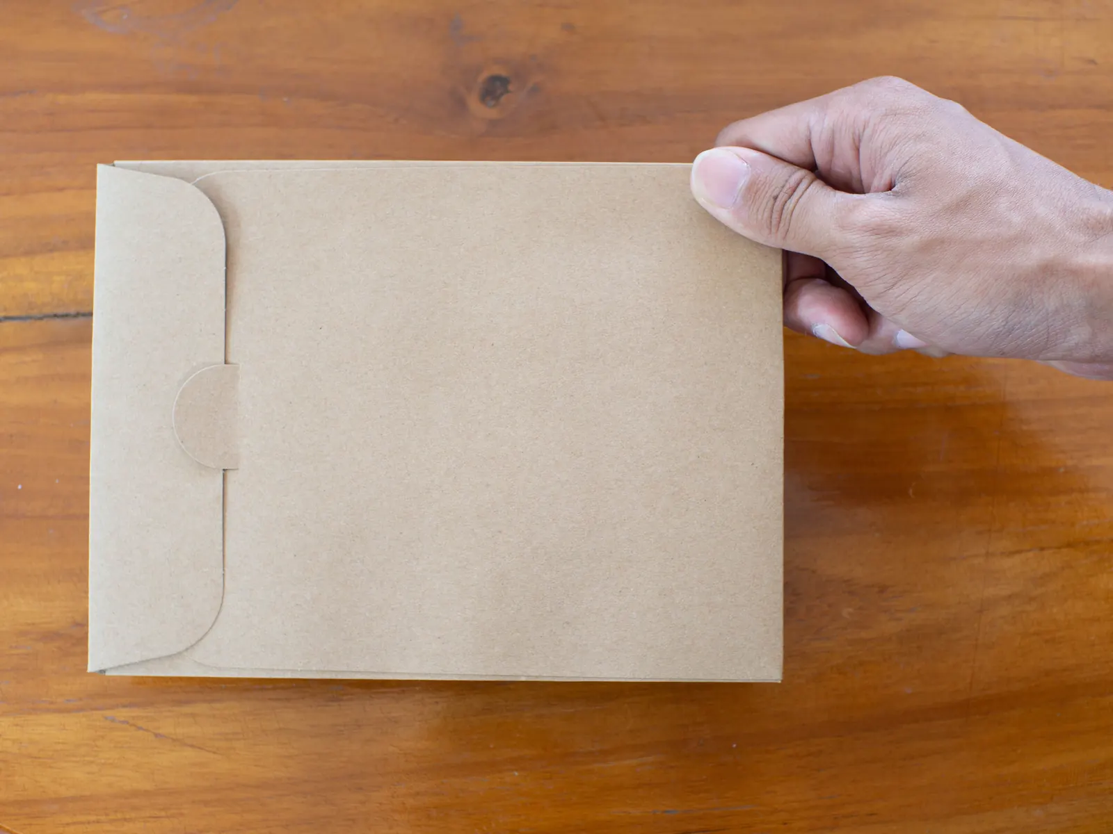 A man's hand slides a plain kraft document envelope across a warm wooden desk