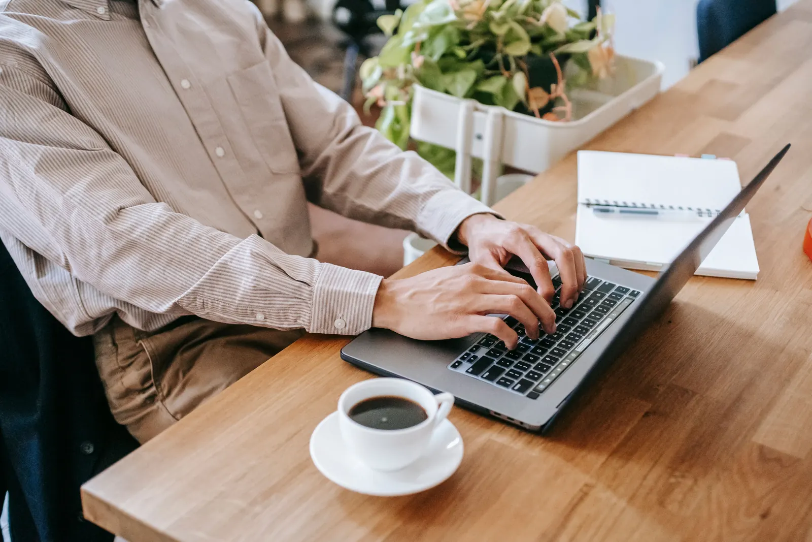 A man in a striped shirt types on a laptop at a wooden desk beside a cup of coffee