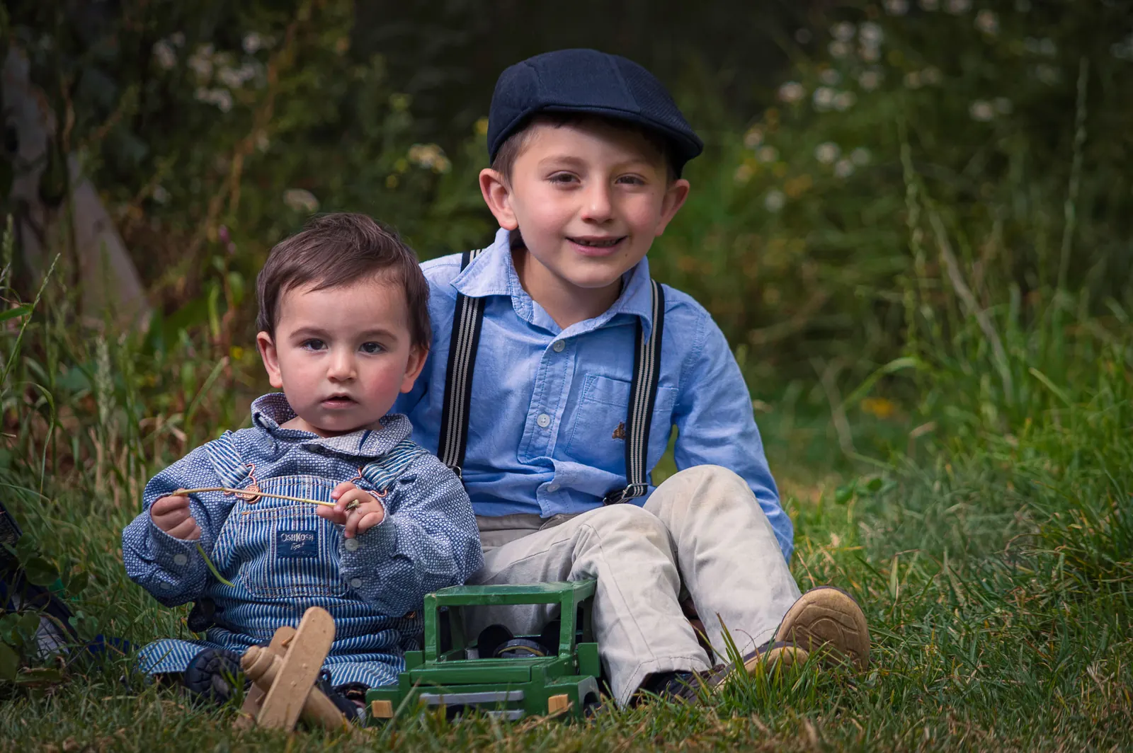 Two young brothers in blue sit together on the grass with a wooden toy truck