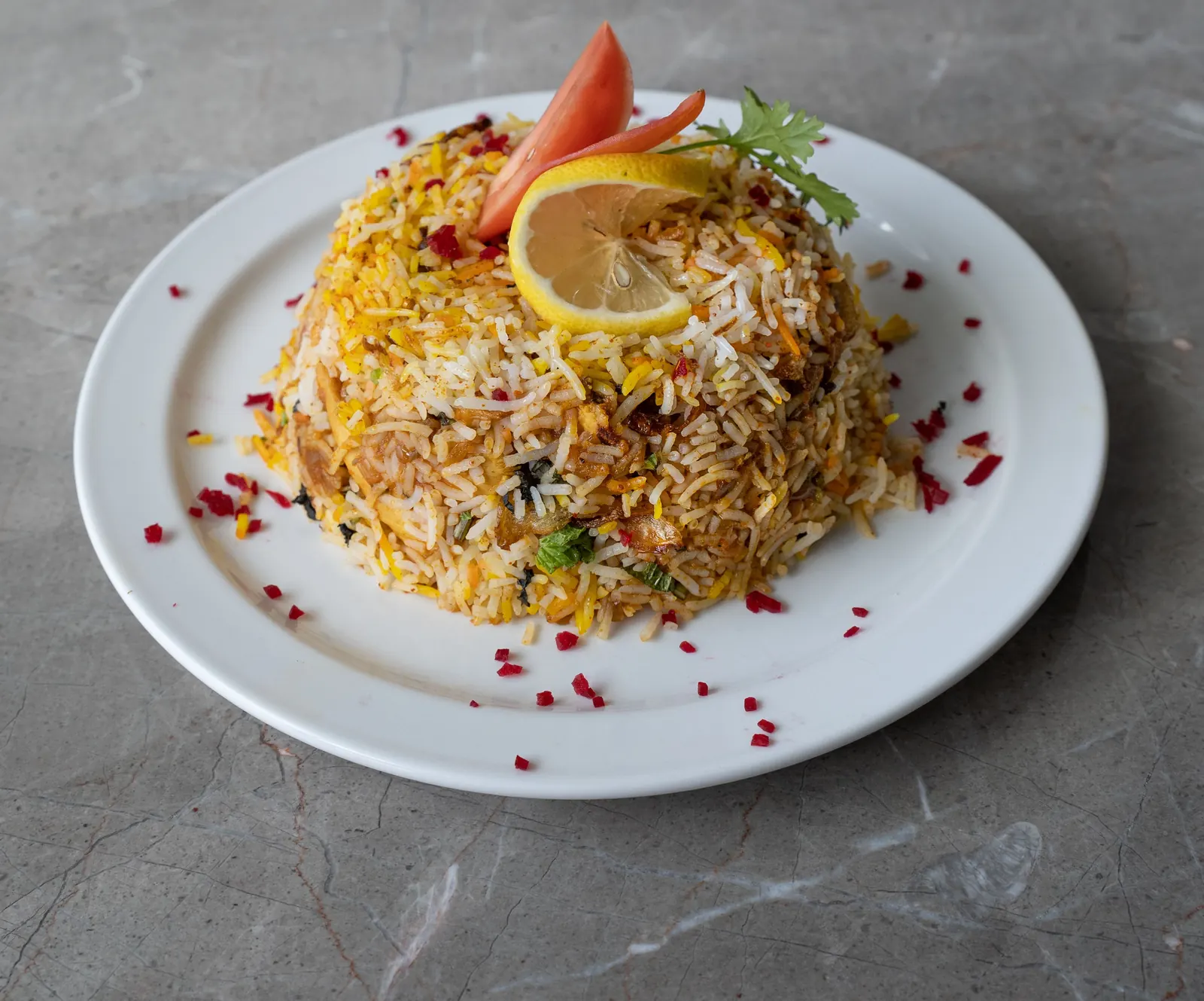 A plate of yellow and white rice biryani topped with lemon and tomato on a grey marble table