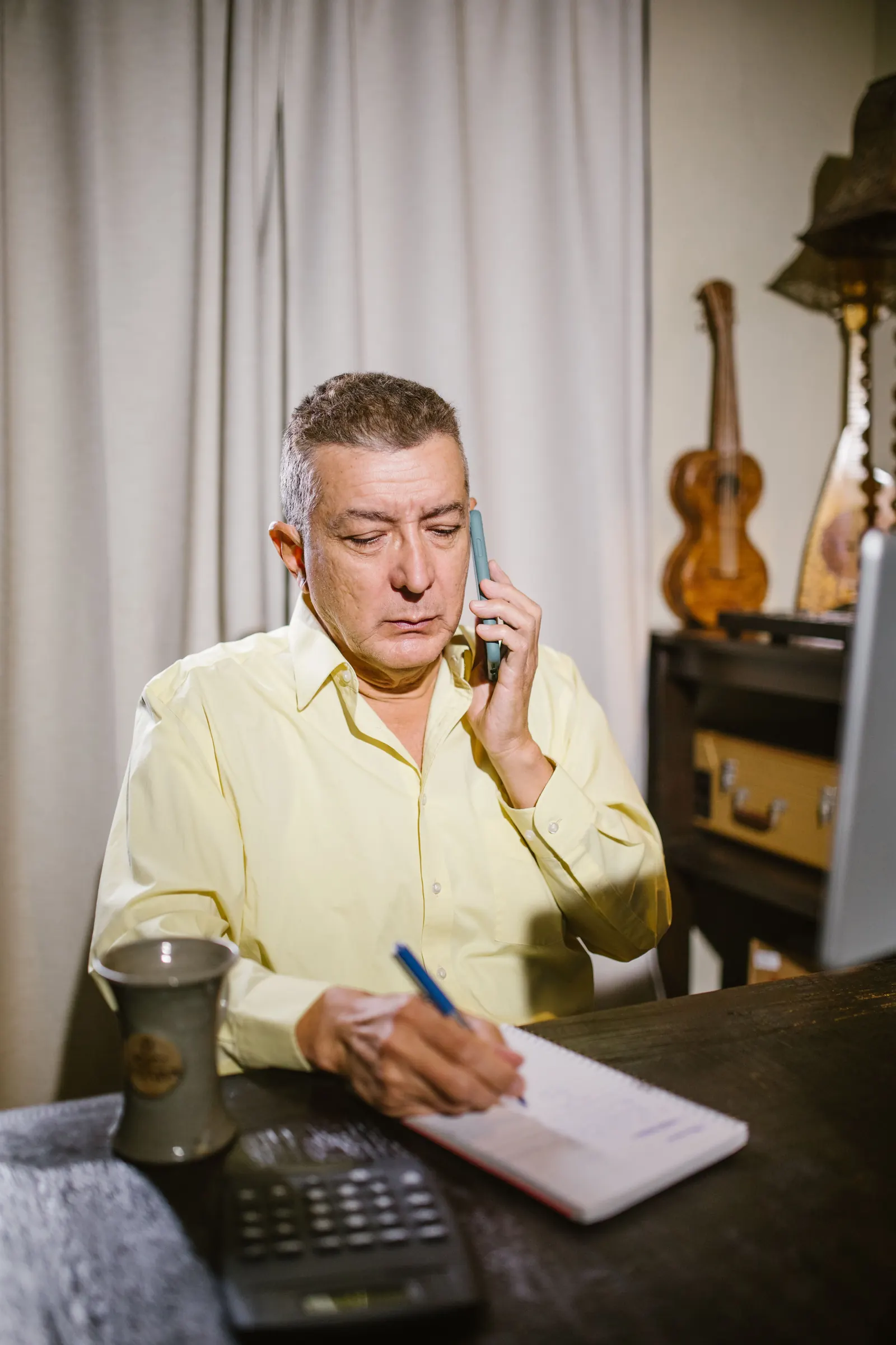 A grey-haired man in a pale yellow shirt talks on his phone while noting details in a notepad at a dark wooden desk