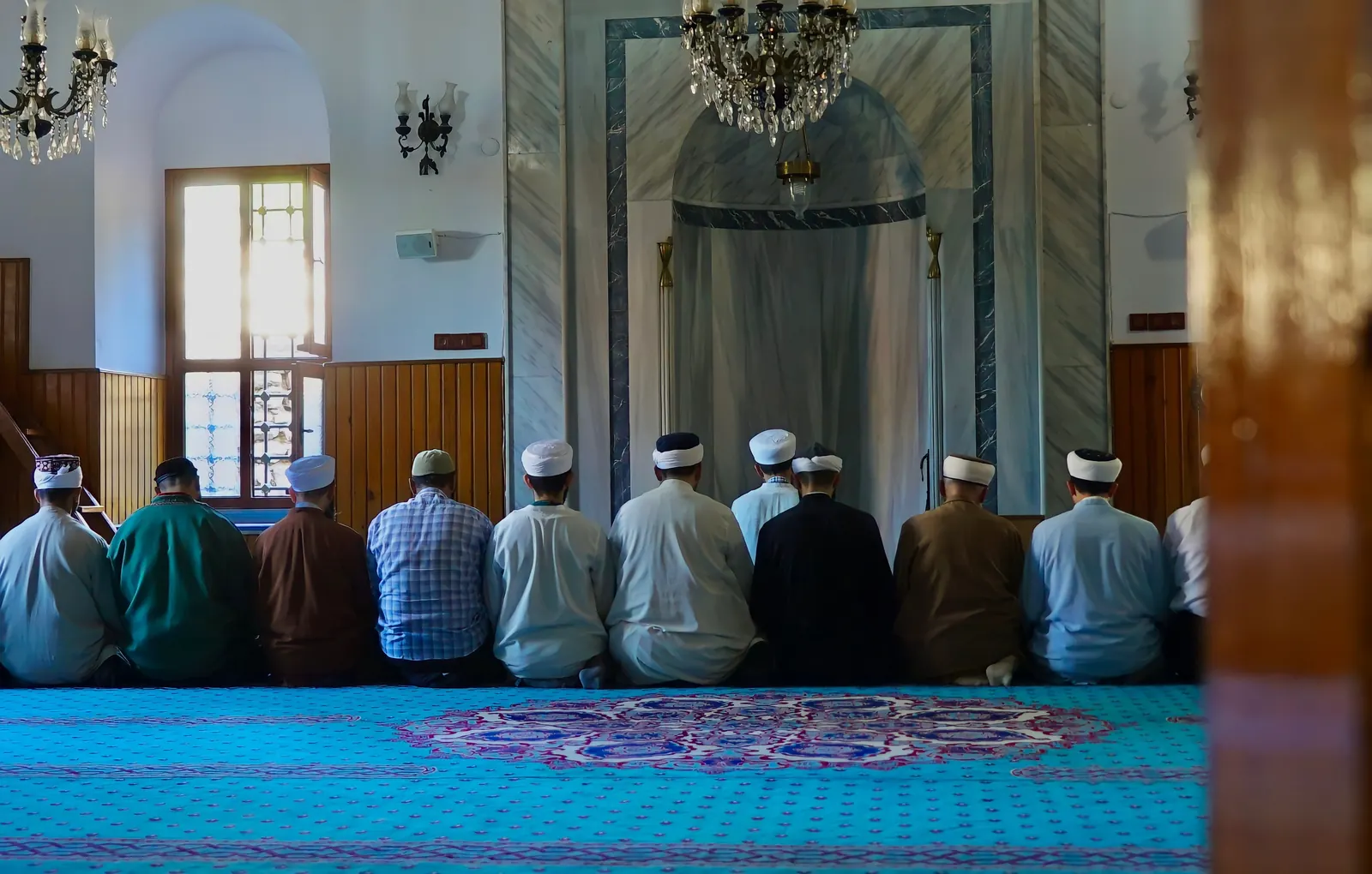 Men in prayer caps kneel in a row on a turquoise carpet in a whitewashed mosque with a chandelier