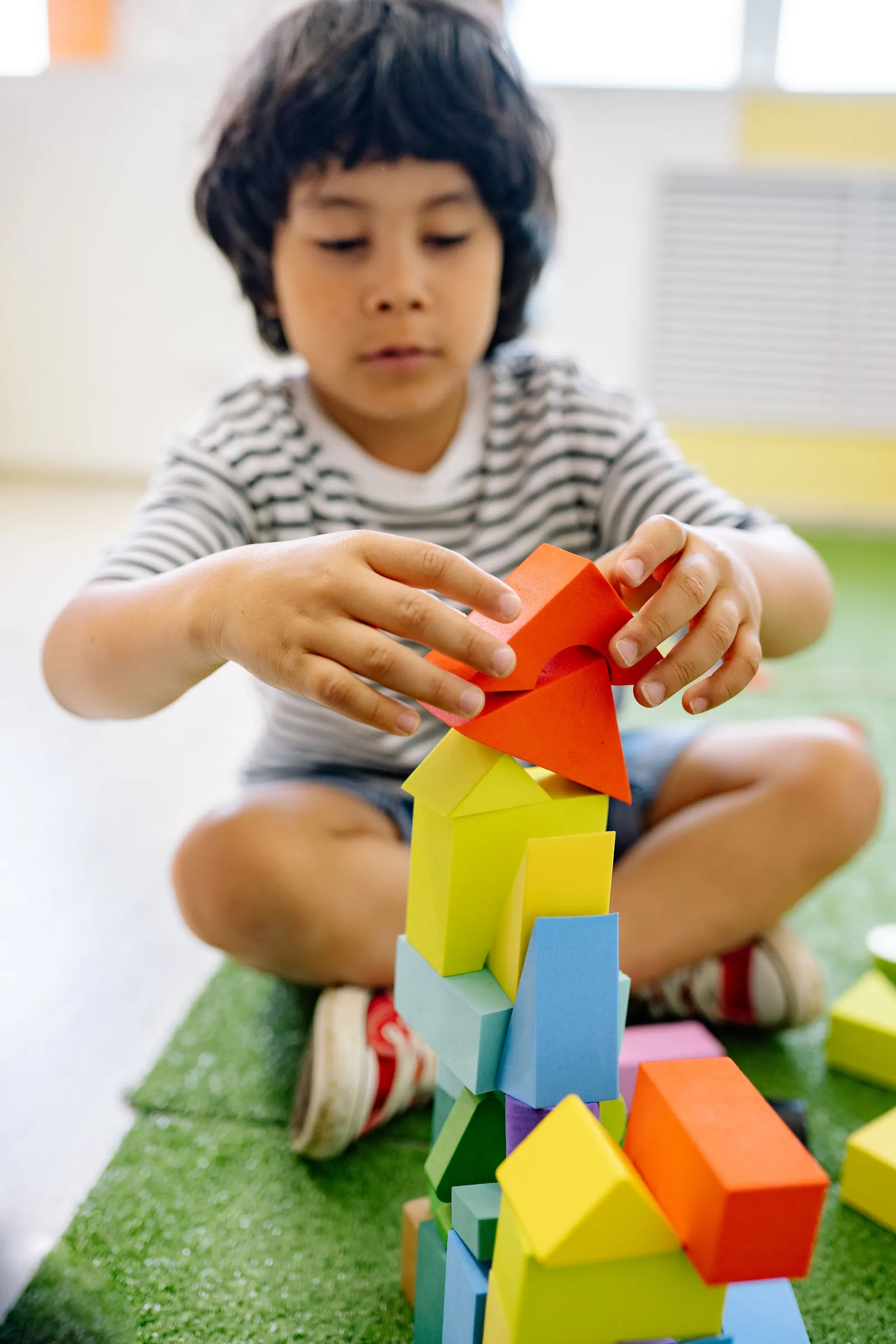 A young boy sits on green matting building a tower of bright foam blocks with both hands