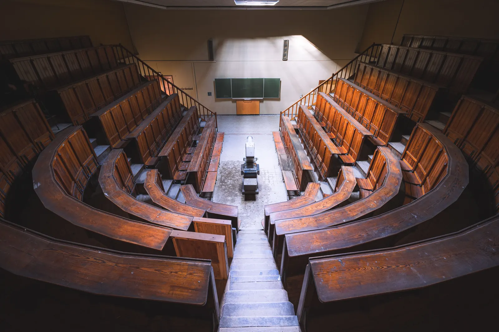An empty tiered lecture hall of curved wooden benches seen from the top, a blank chalkboard below