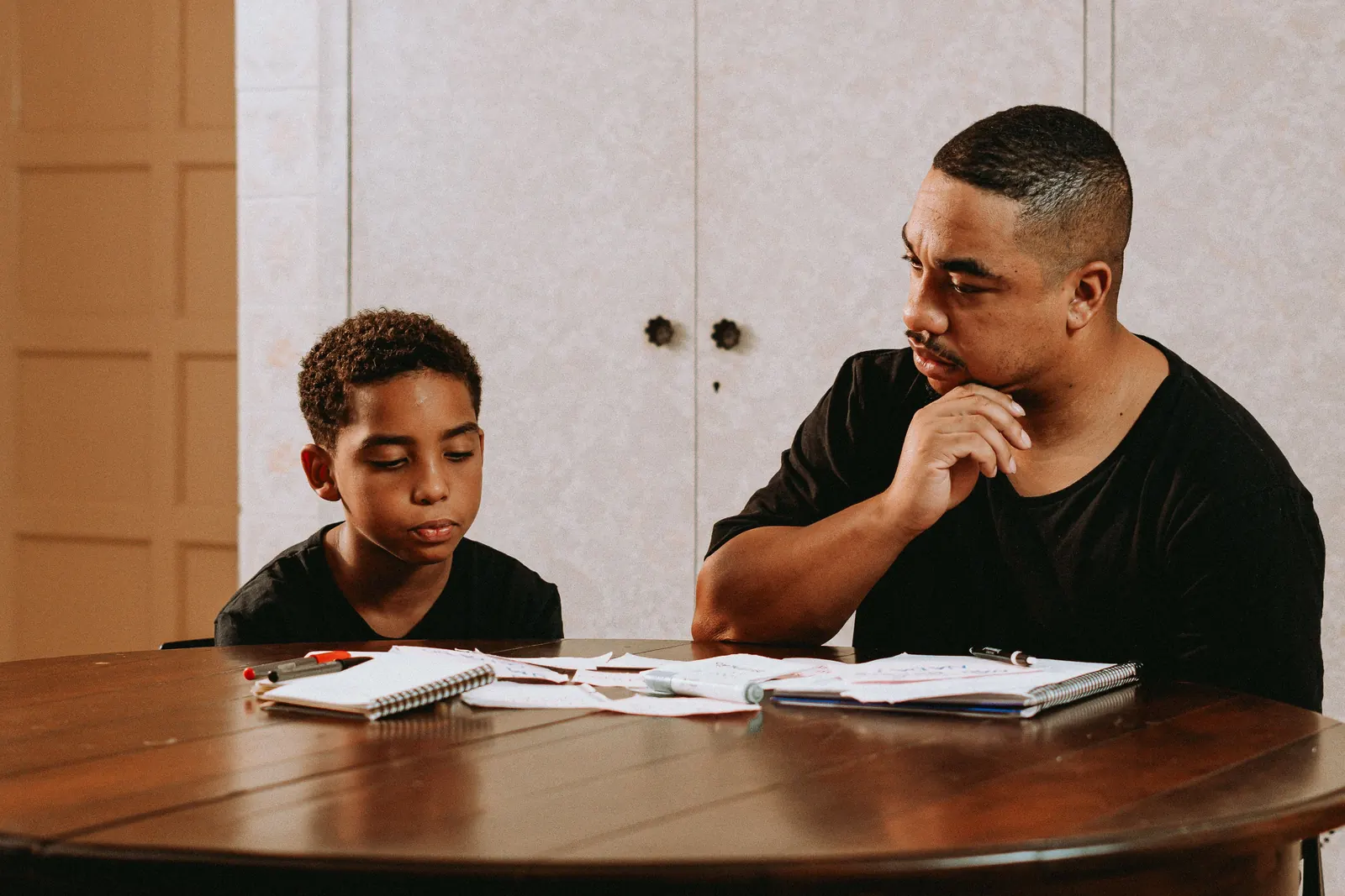 A man sits at a wooden table beside a boy and watches him work through his notebook
