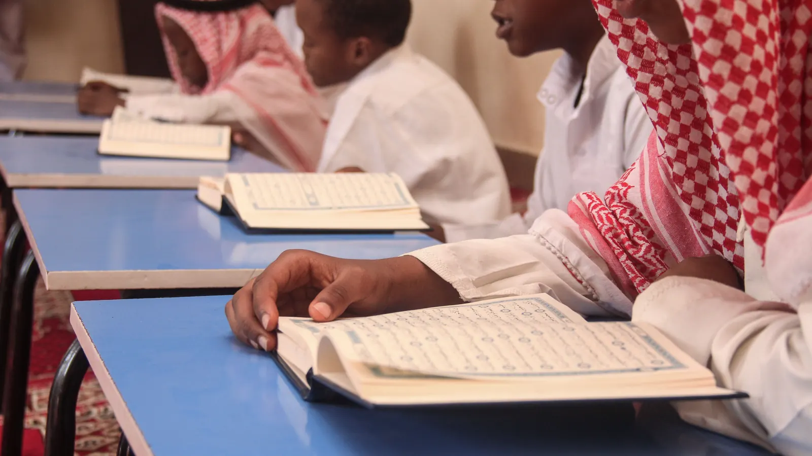 Boys in white thobes and red-checked headscarves read open Qur'ans at blue classroom desks