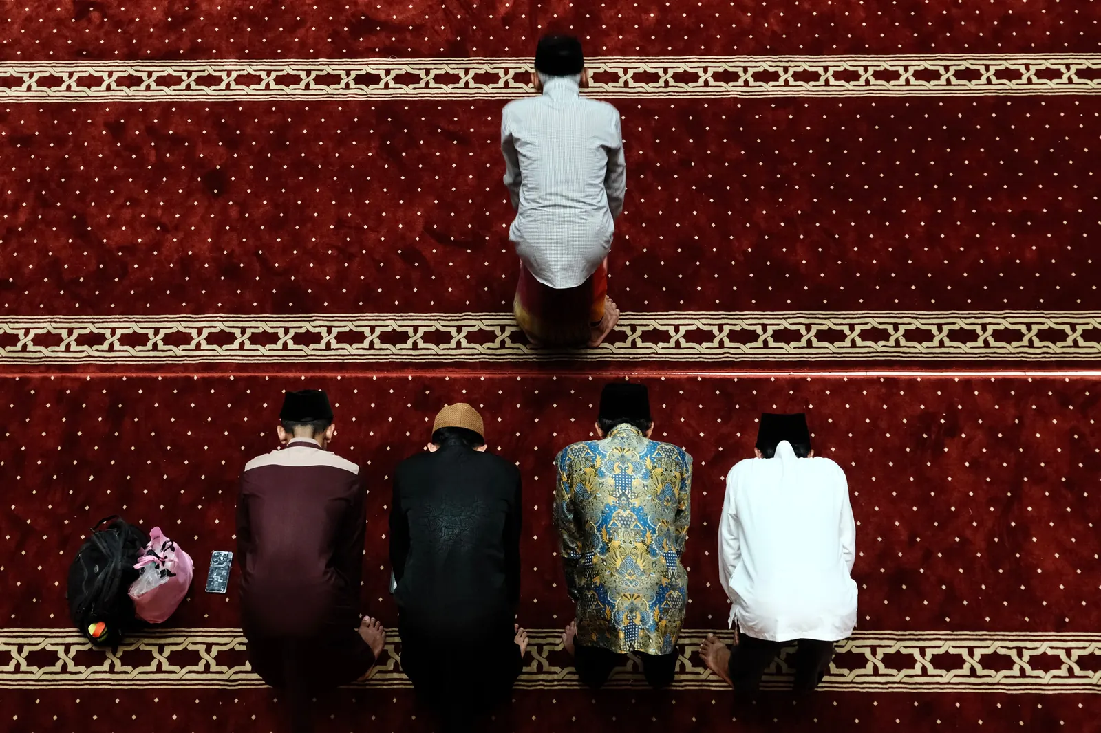 Seen from above, a row of men and boys kneel on a red prayer carpet behind the one leading the prayer
