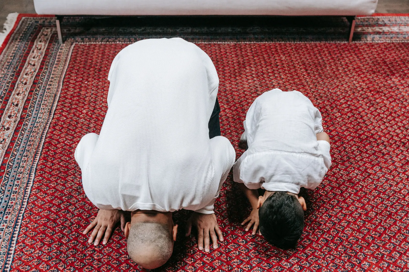 A father and his young son, both in white, bow side by side in sujood on a red rug