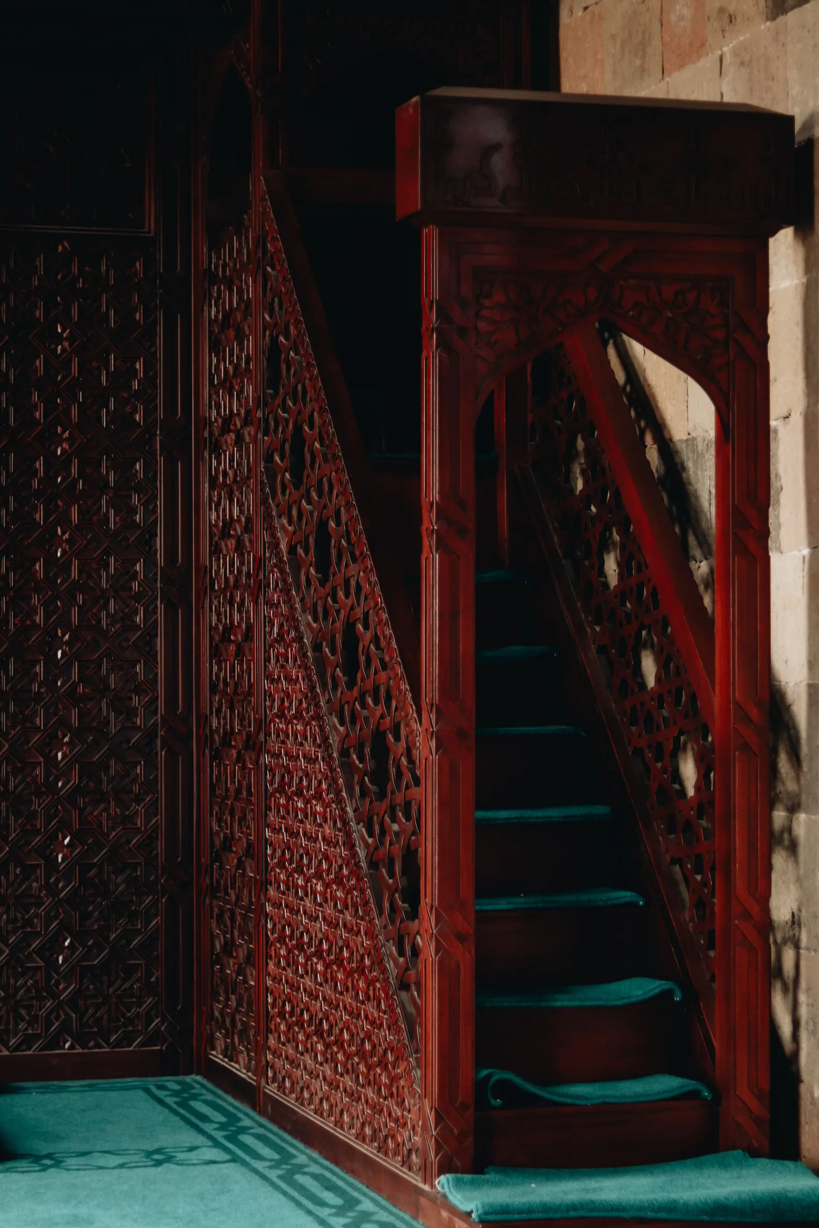 A carved red-wood minbar with lattice sides and teal carpeted steps rises beside a stone wall