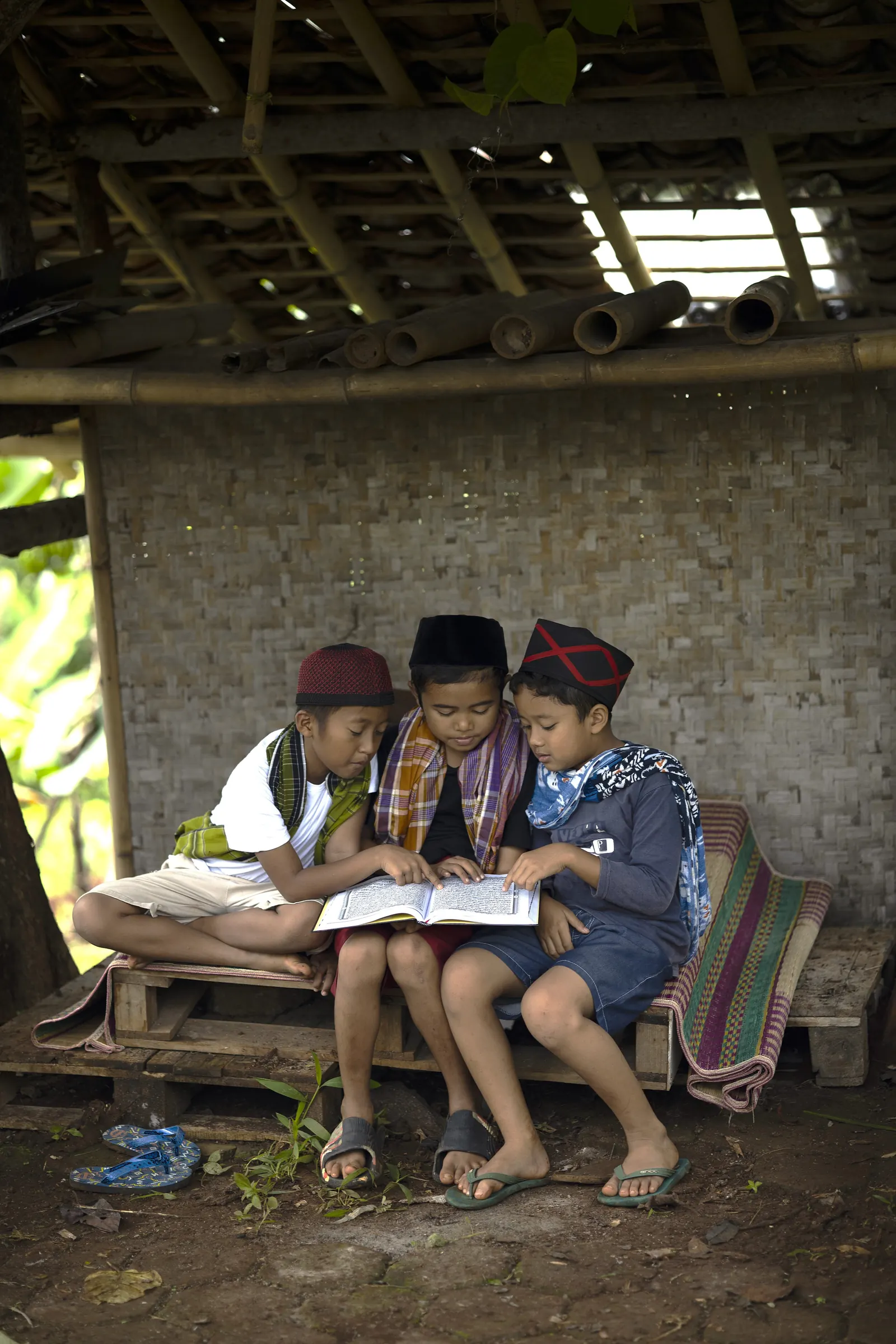 Three boys in caps and scarves sit close on a bench under a bamboo shelter, reading one book together