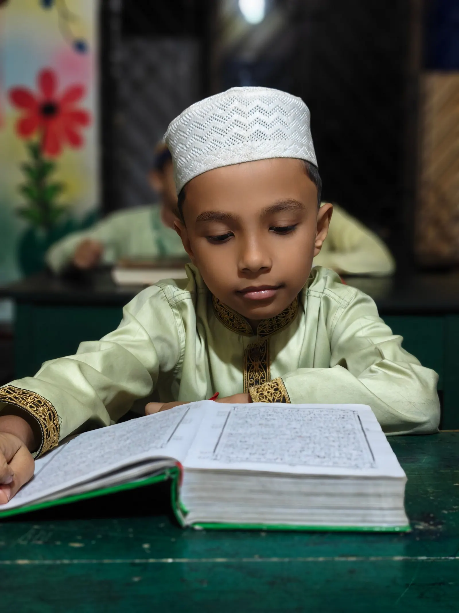 A boy in a white kufi and embroidered kurta reads a large open book at a green school desk