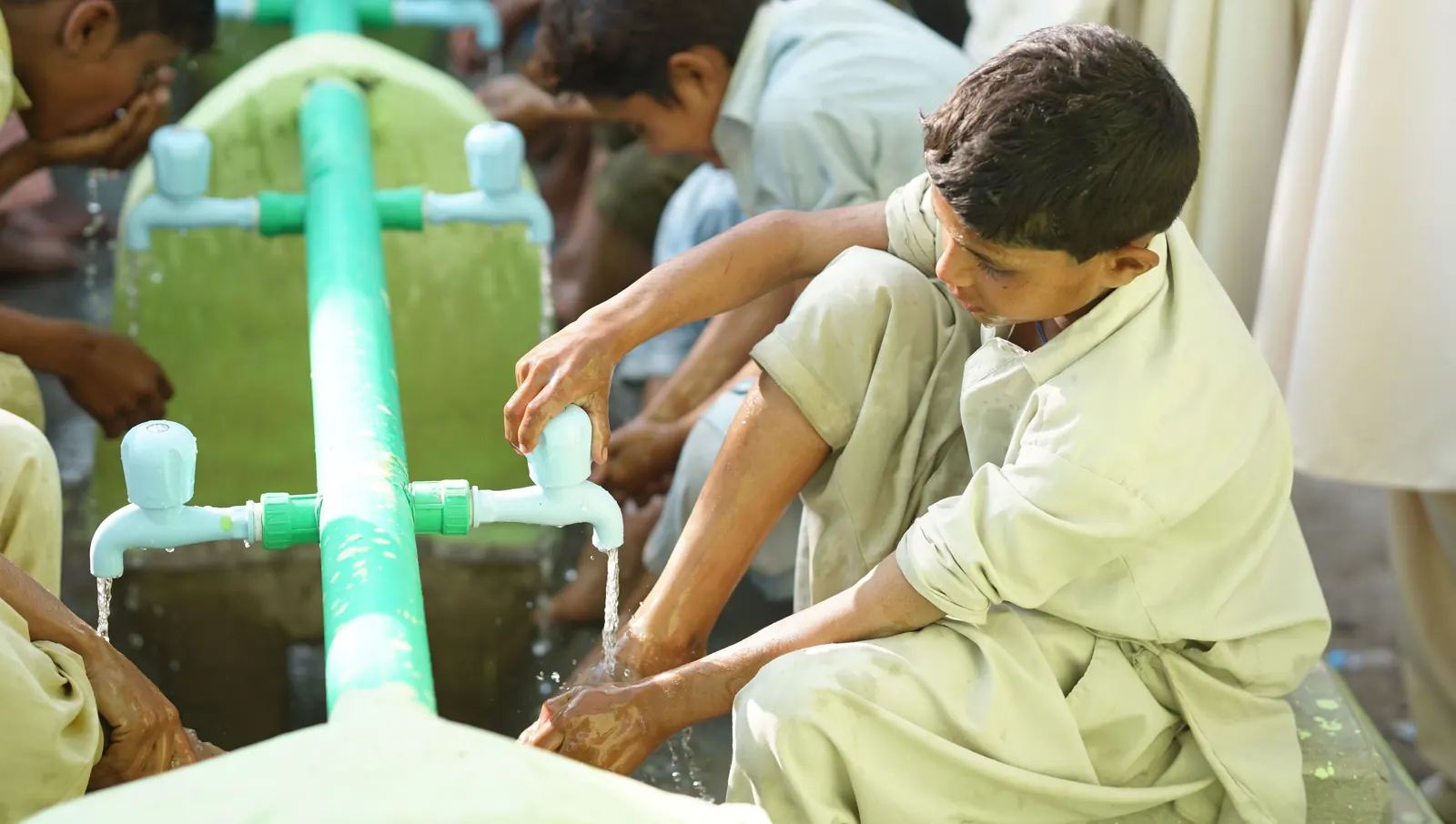 Boys in shalwar kameez make wudu at a row of taps along a green pipe outdoors
