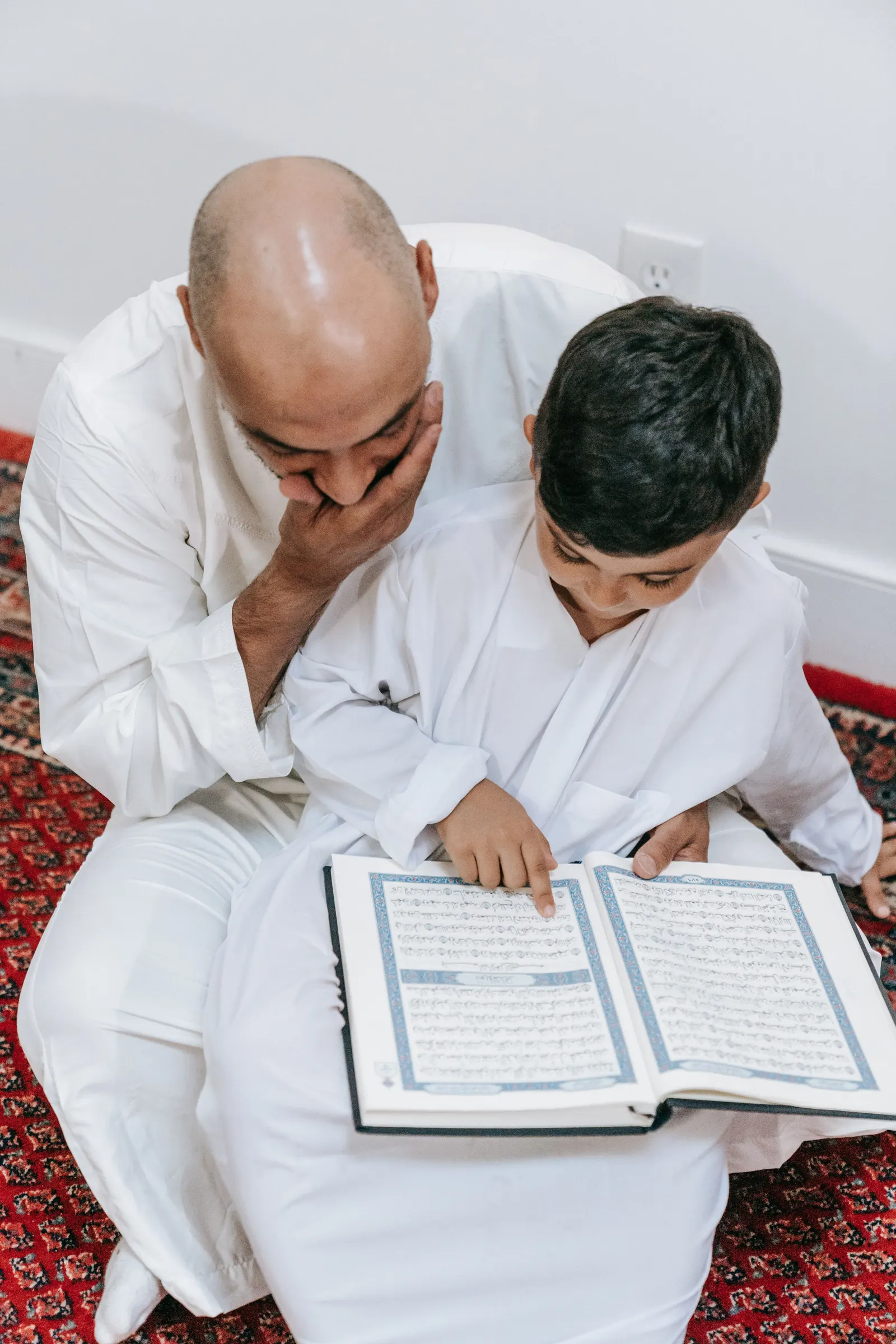 A man in a white thobe sits behind a boy, following as the boy points along the lines of an open Qur'an
