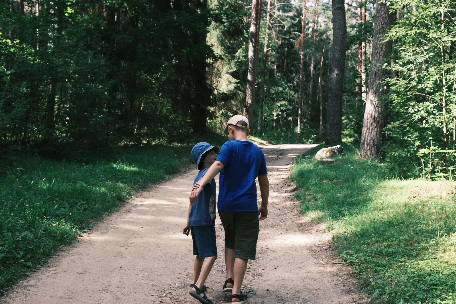 An older boy walks along a forest path with his arm around a younger boy who looks up at him