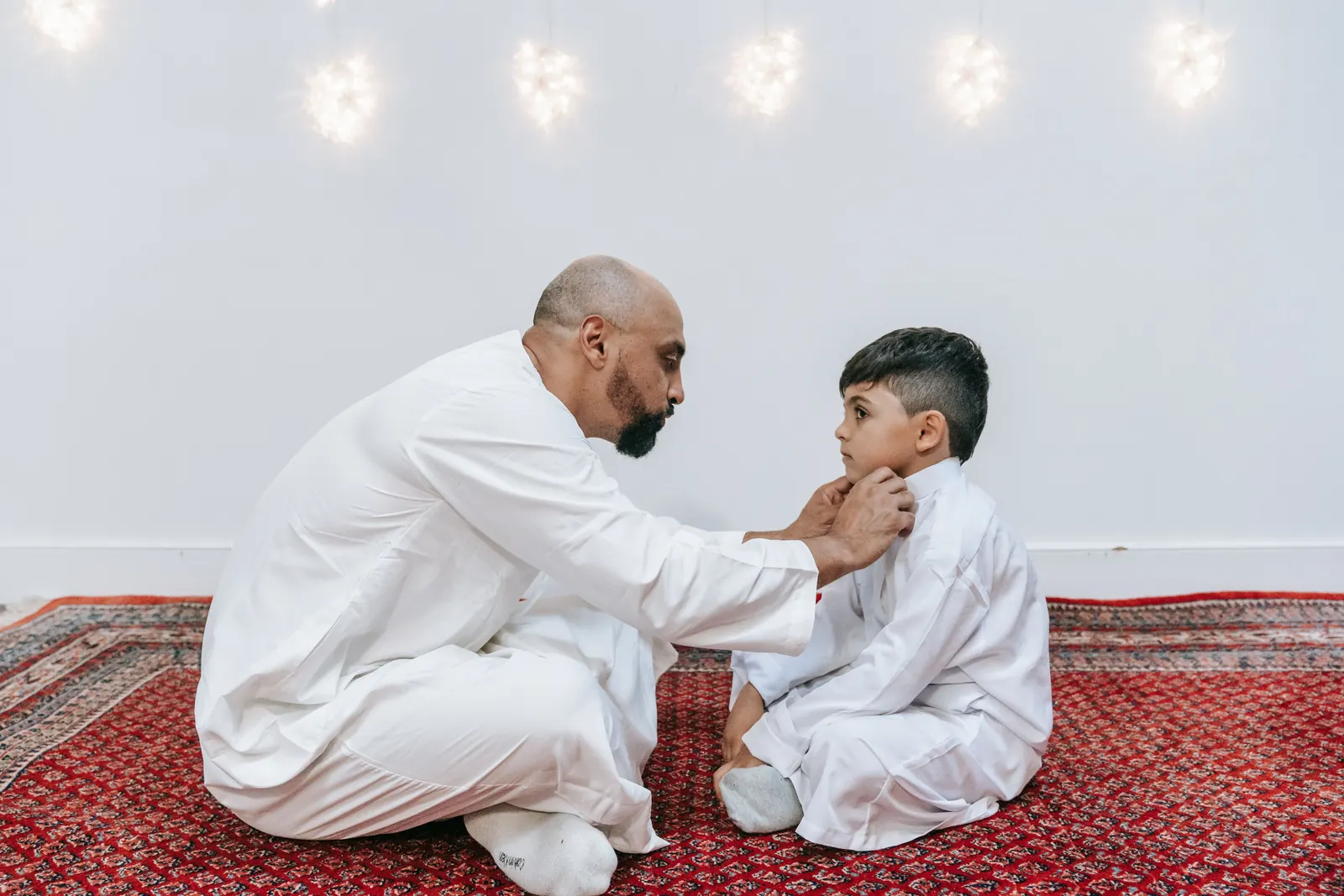 A father in a white thobe kneels on a red rug to straighten his young son's collar before prayer