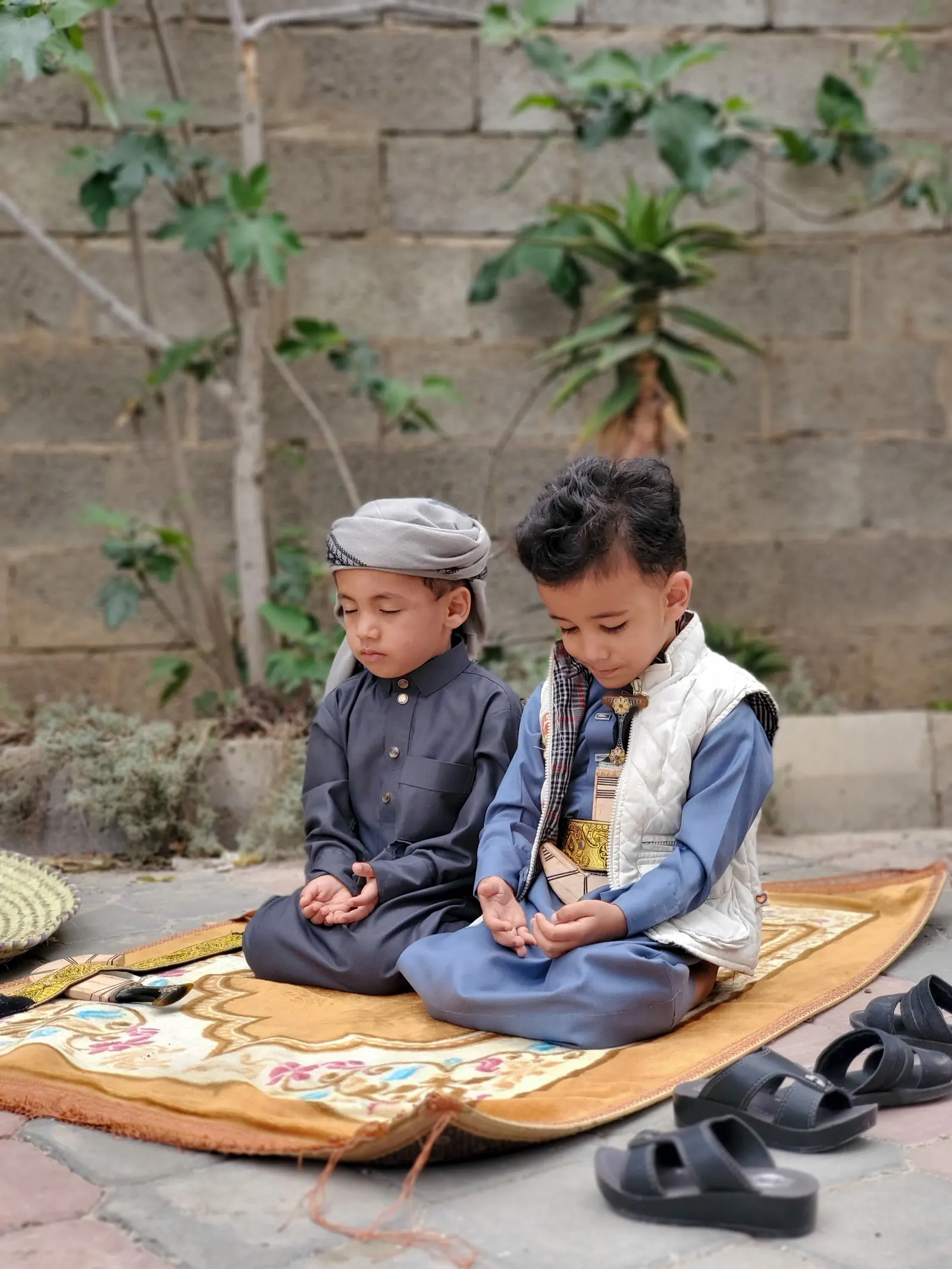 Two small boys in thobes sit on a prayer mat outdoors, eyes lowered and hands raised in dua