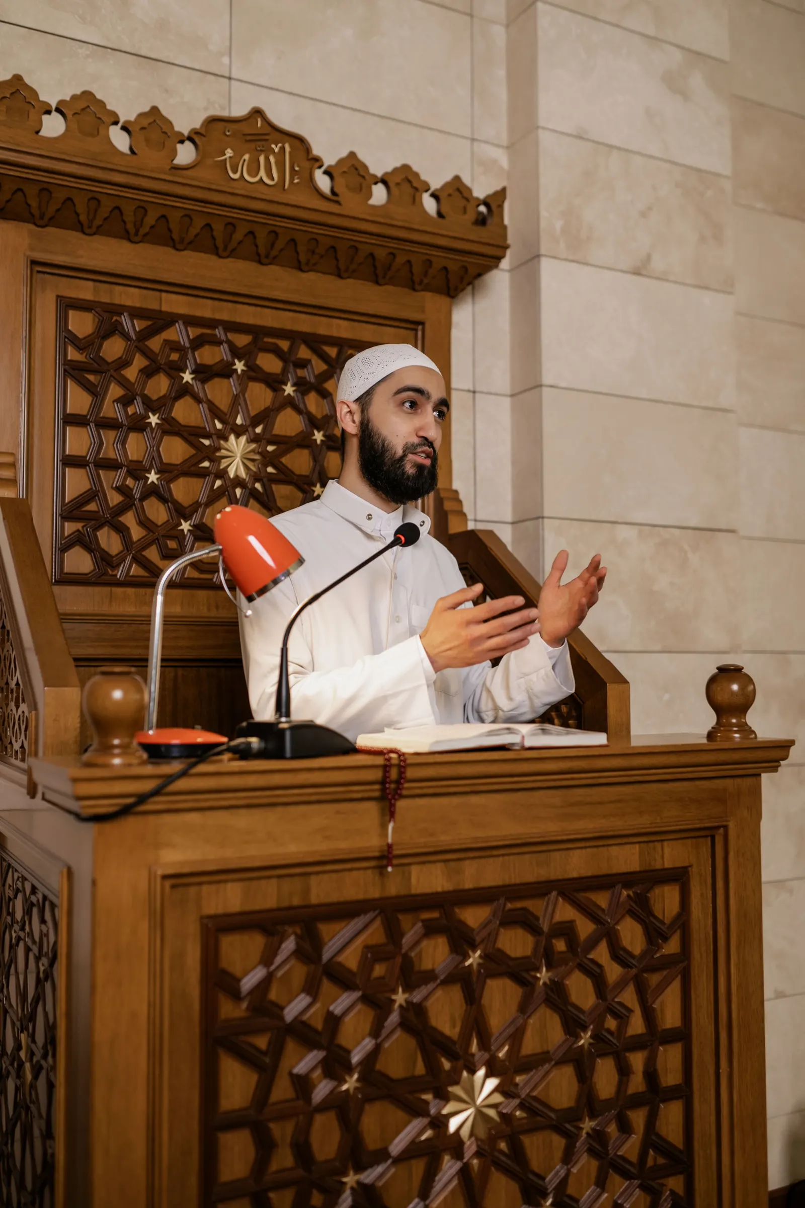 A young imam in a white kufi speaks from a carved wooden minbar lectern with a microphone