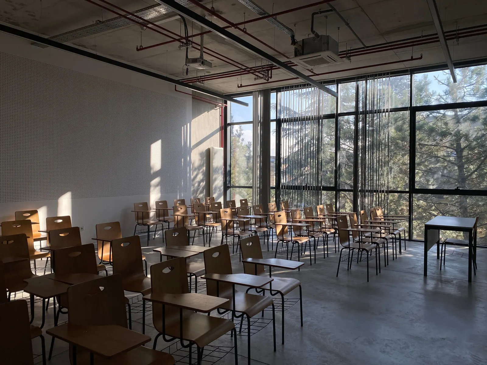 An empty classroom of wooden tablet-arm chairs lit by low sun through tall windows onto pine trees